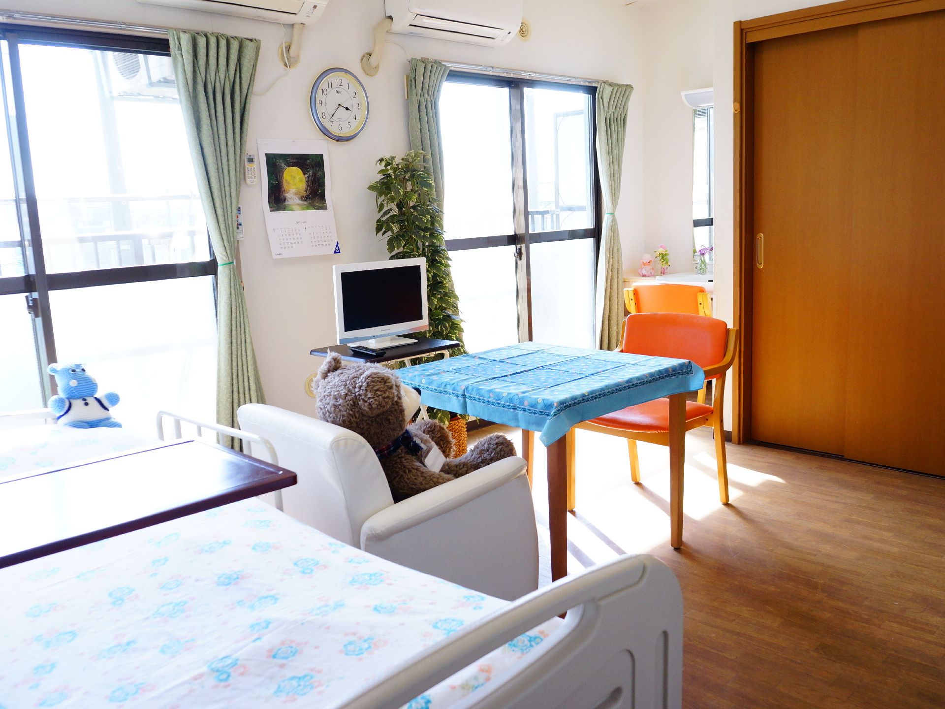 部屋には大きな窓が2つあり、太陽の光がいっぱいに差し込む。イスとテーブルの前にはテレビが置かれ、壁には時計が掛かっている。