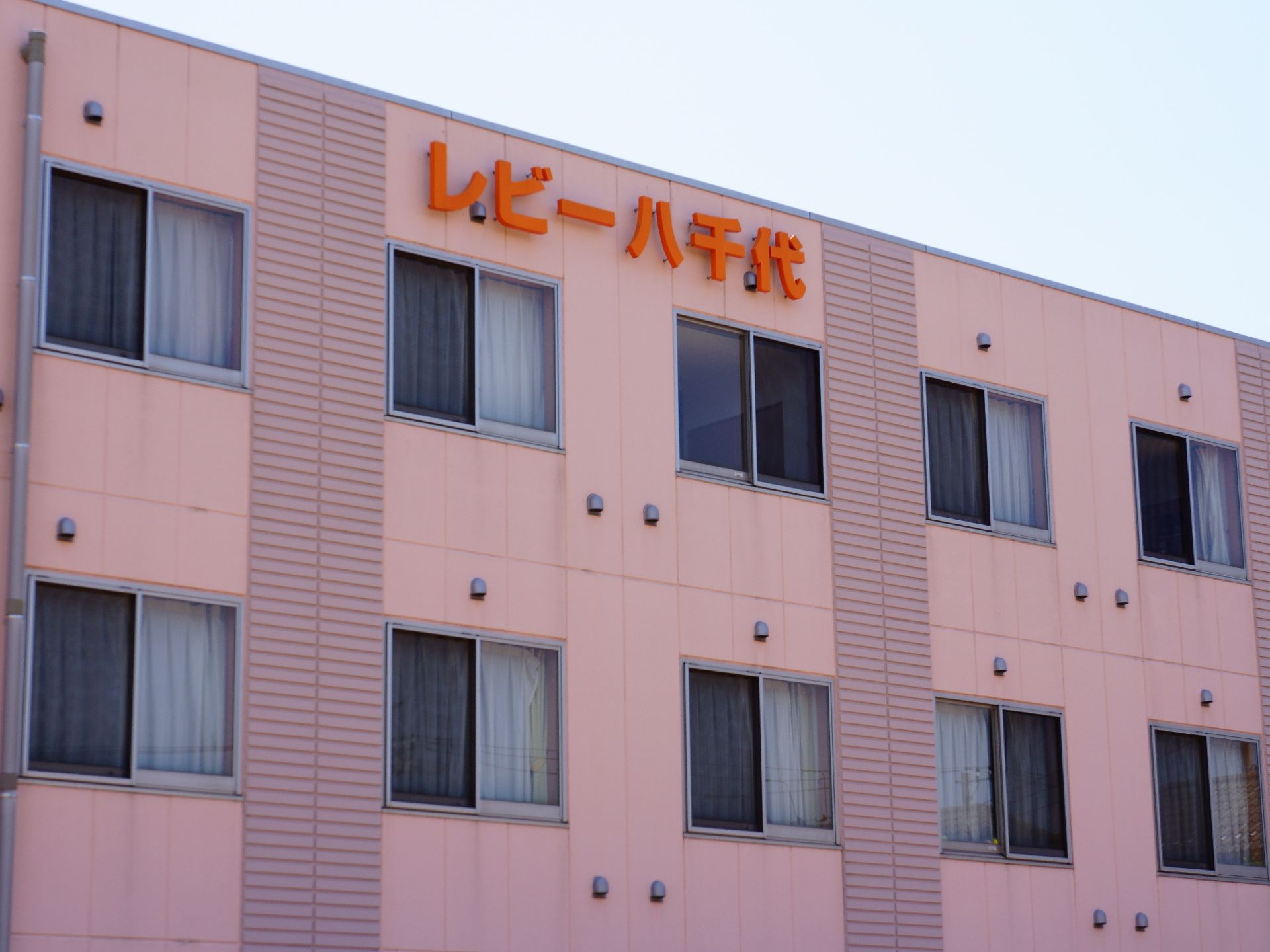 当施設の外観の様子。外観は淡いピンク色でまとめられている。施設上部には、くっきりと目立つように施設名がオレンジで掲示されている。