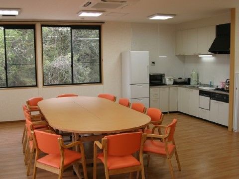 大きな長丸形のテーブルに、オレンジの座面の手すりのついた椅子がいくつも置かれている。家庭用キッチンでちょっとした料理もできる。