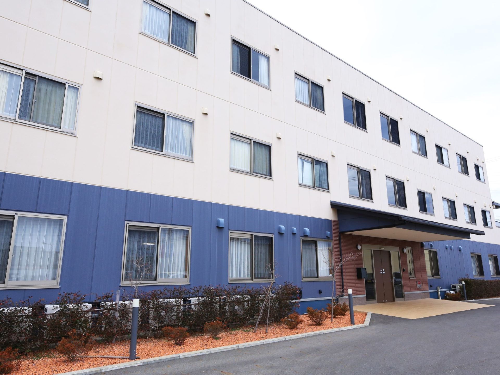 正面から見た建物の玄関付近の様子。１階部分の外壁には青い材質が使われており、２階から上は白くなっている。