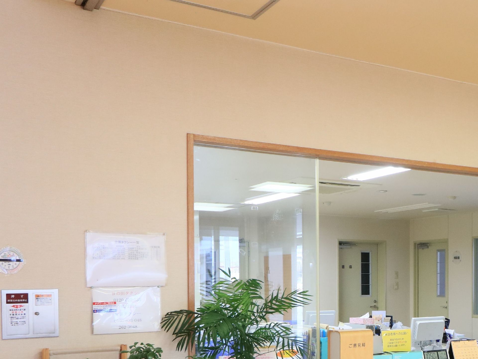 観葉植物の向こう側に受付の窓があり、書類やパソコンが置いてある部屋がある。壁に数枚の紙が貼られ、ご意見箱が設置されている。
