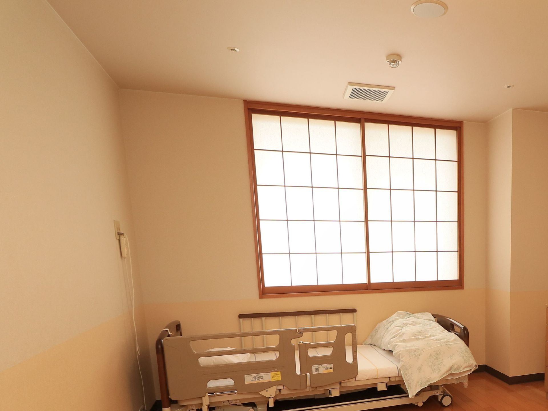 白いクロスが張られた壁とフローリングの床の室内である。障子が取りつけられた腰高窓の下にベッドが置いてある。