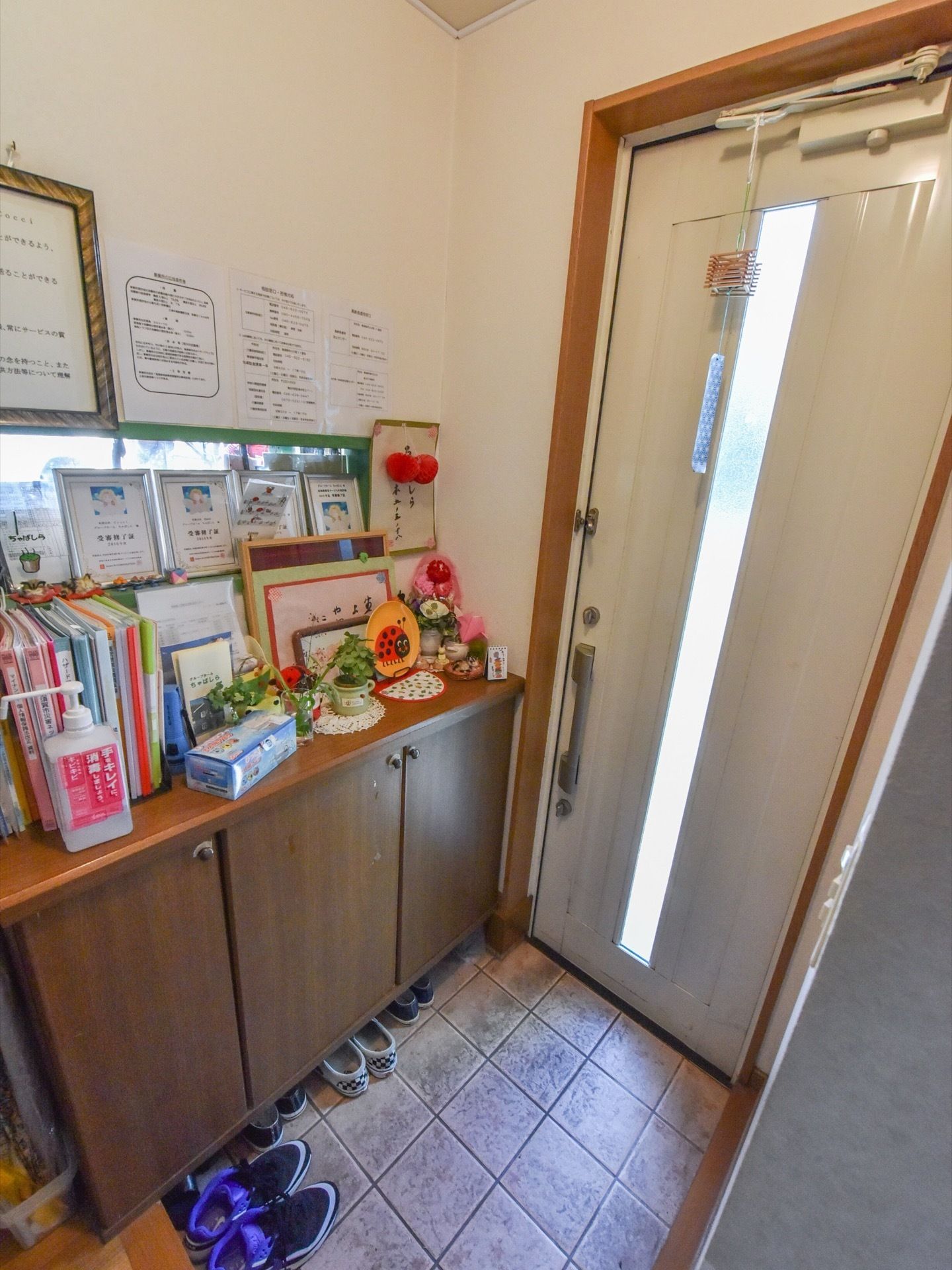 玄関の扉には鈴が取り付けられている。下駄箱の上には何枚かの受審終了証が並べられて掲げられている。植物も置かれている。