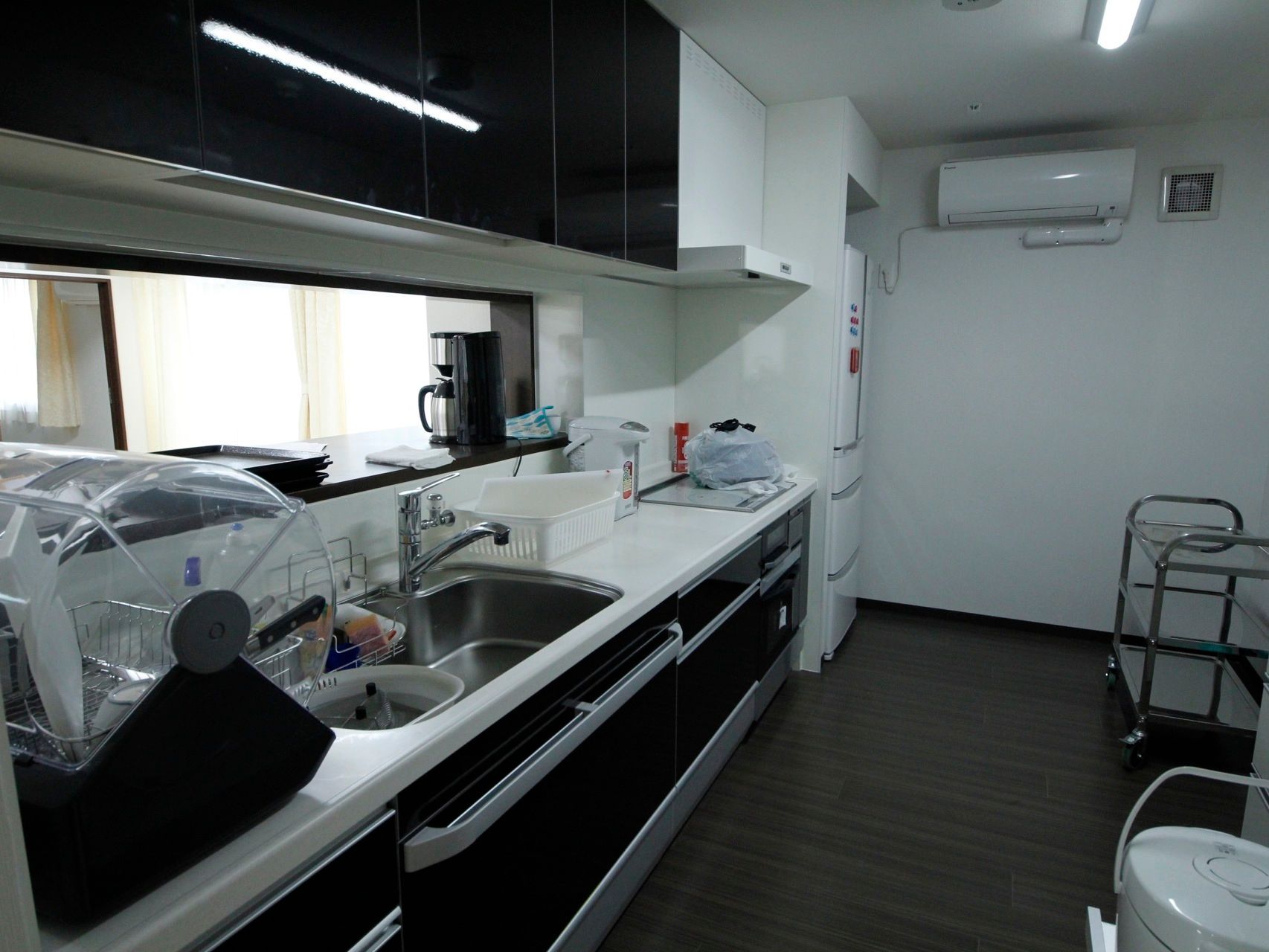 動きやすい広さのキッチンである。シンクや作業台の上には、食洗機やポットが置かれており、奥のスペースに冷蔵庫が設置されている。