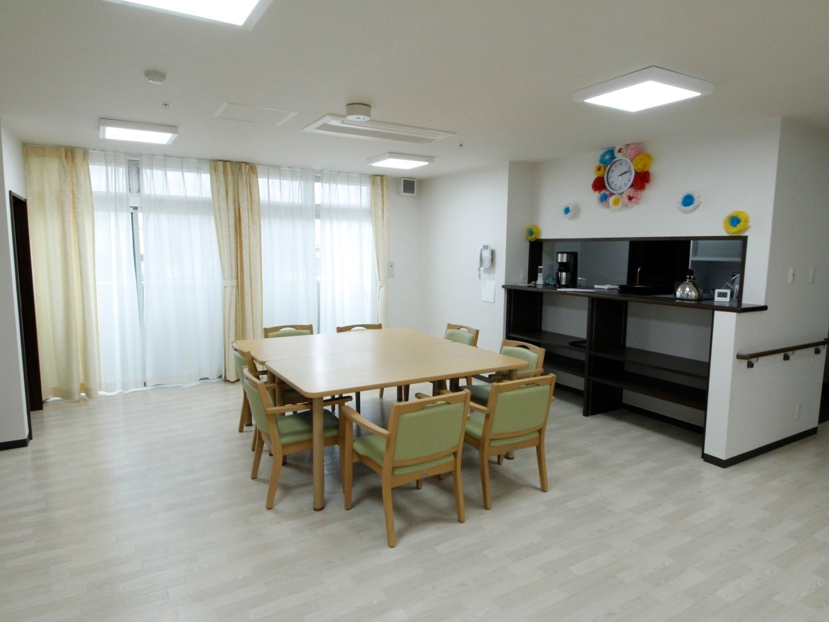 開放的で明るい食堂である。８人掛けテーブルが置かれている。右側にはキッチンがある。手すりや時計、電話機が壁に設置されている。