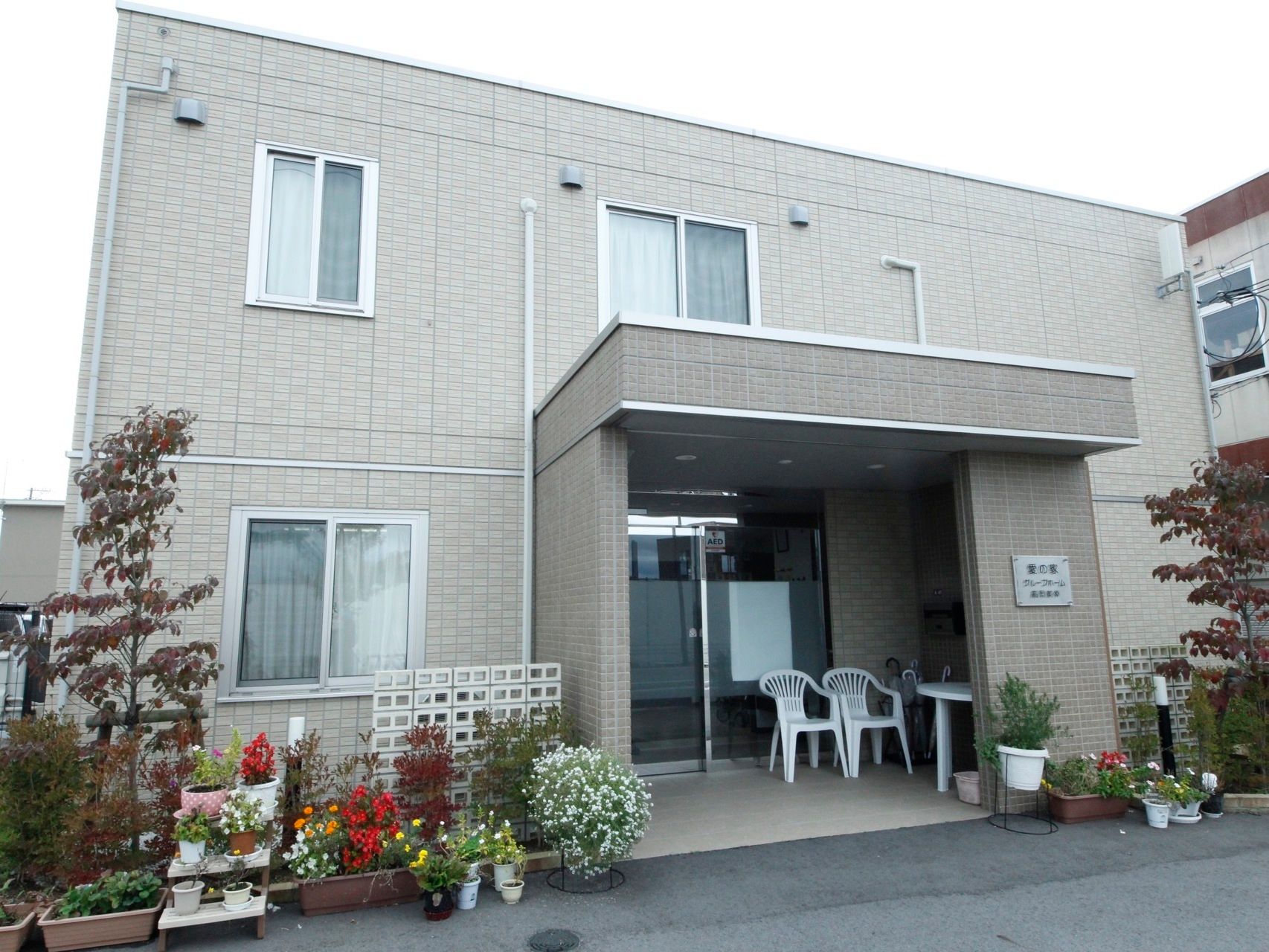 ２階建ての施設である。壁際には、色々な種類の花が植えられたプランターが置かれている。玄関ポーチに白いテーブルと椅子が２脚置かれている。