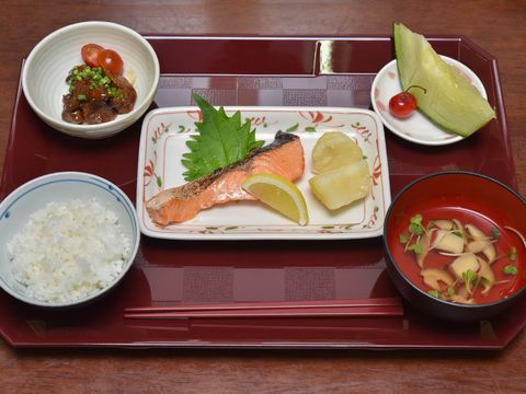 トレーの上に、ご飯、主菜のさけの塩焼きと、肉団子がある。きのこの吸い物とデザートのレモンが添えられている。