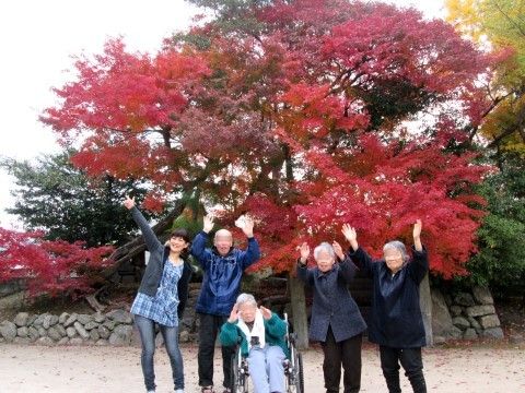 赤く色づいた紅葉の下、手を上げて記念撮影をする入居者たちと職員