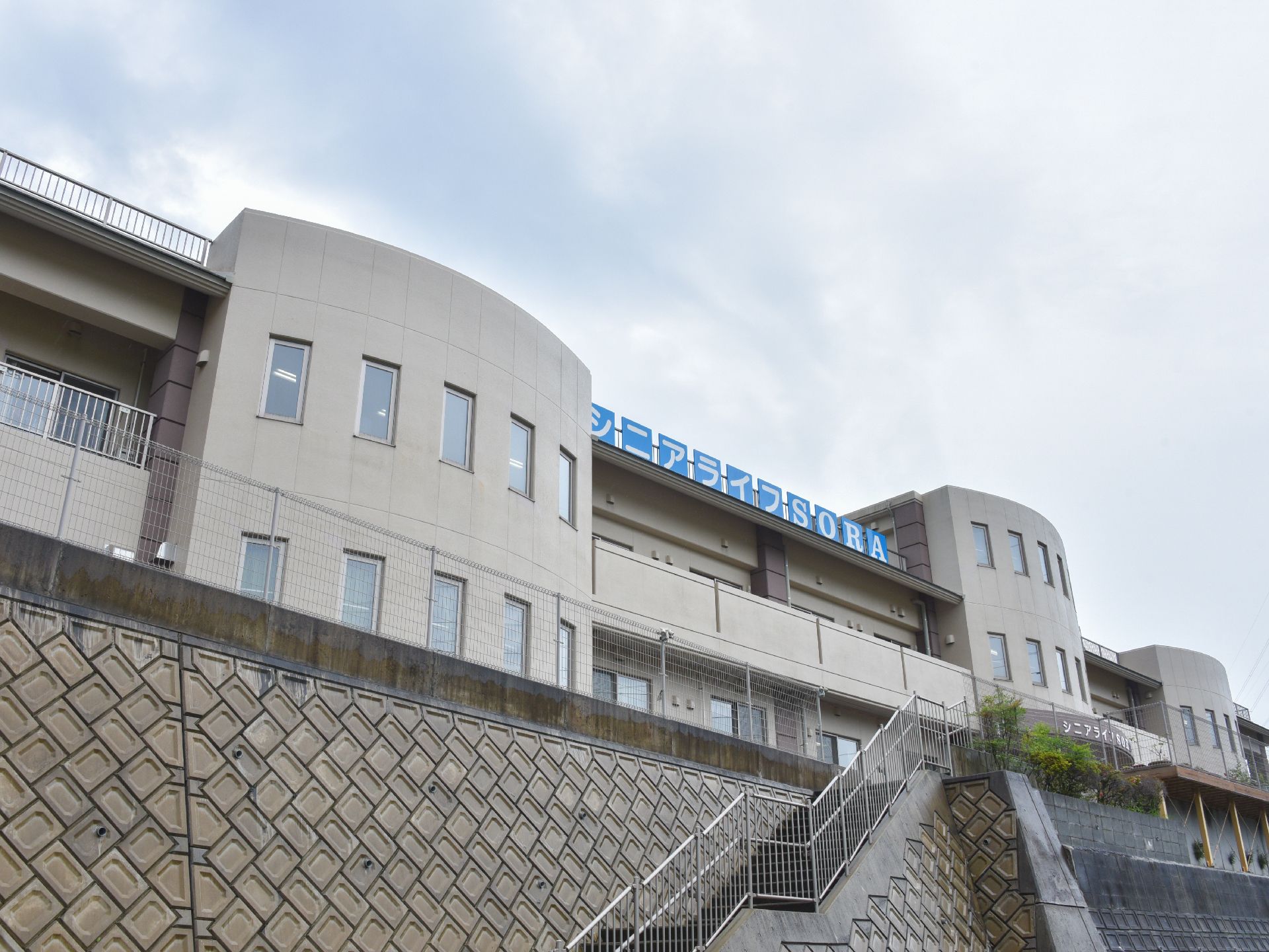 高台に建っている施設は、一部は3階建てであるが大半は2階建てである。外壁はベージュ色になっており、ベランダや窓が見える。