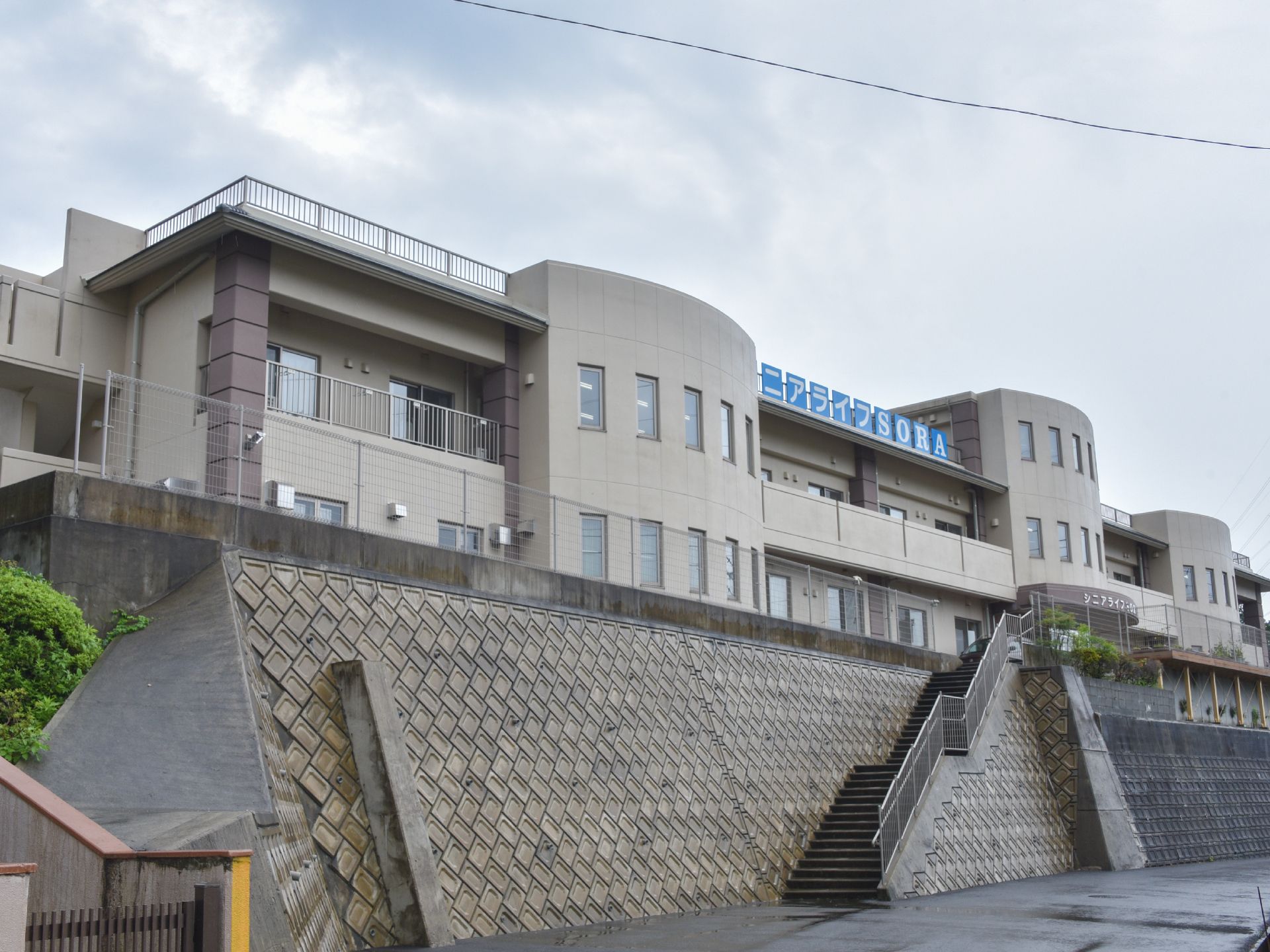 手前には道路があり、施設は高台に建っているため、階段が用意されている。建物は一部は3階だが大半は2階建てである。