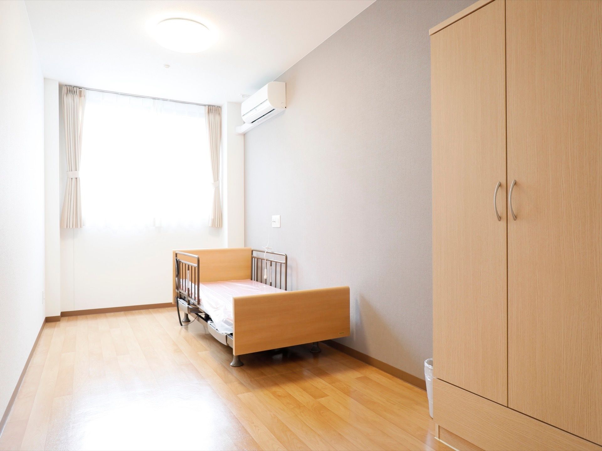 フローリングの部屋に電動ベッドが設置されている。壁にはエアコンがついている。手前には観音開きの戸がついたクローゼットがある。