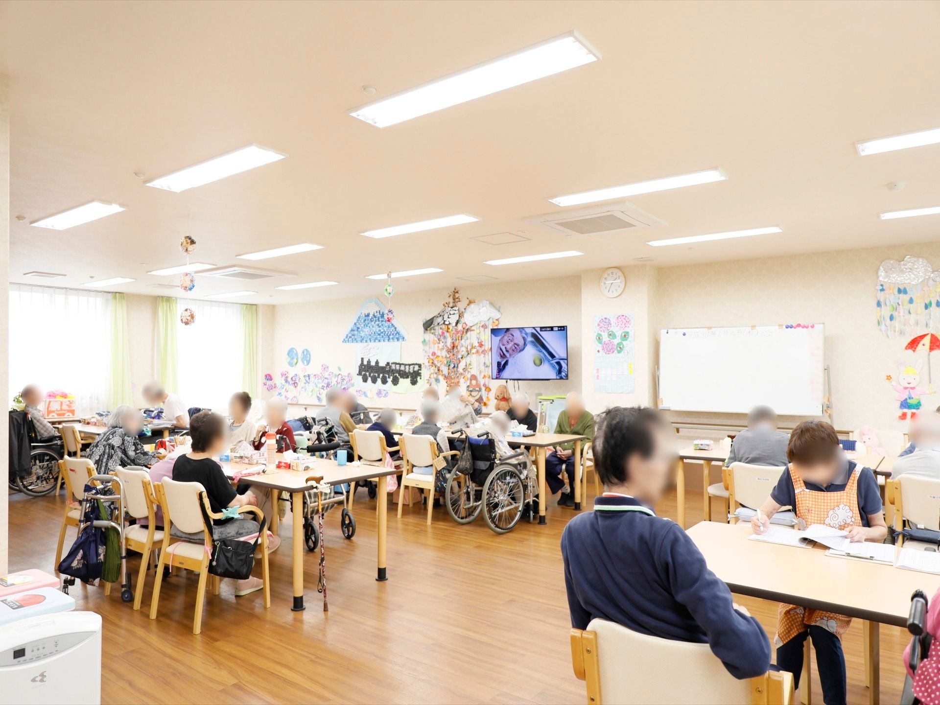 ダイニングテーブルがたくさん並び、入居者様が揃って席についている。テレビを観たり、スタッフと作業をしたりしている方もいる。