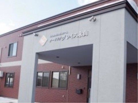 玄関に屋根がついている。グレーの外壁には施設名が書かれており、上部には照明が２つ取り付けられている。