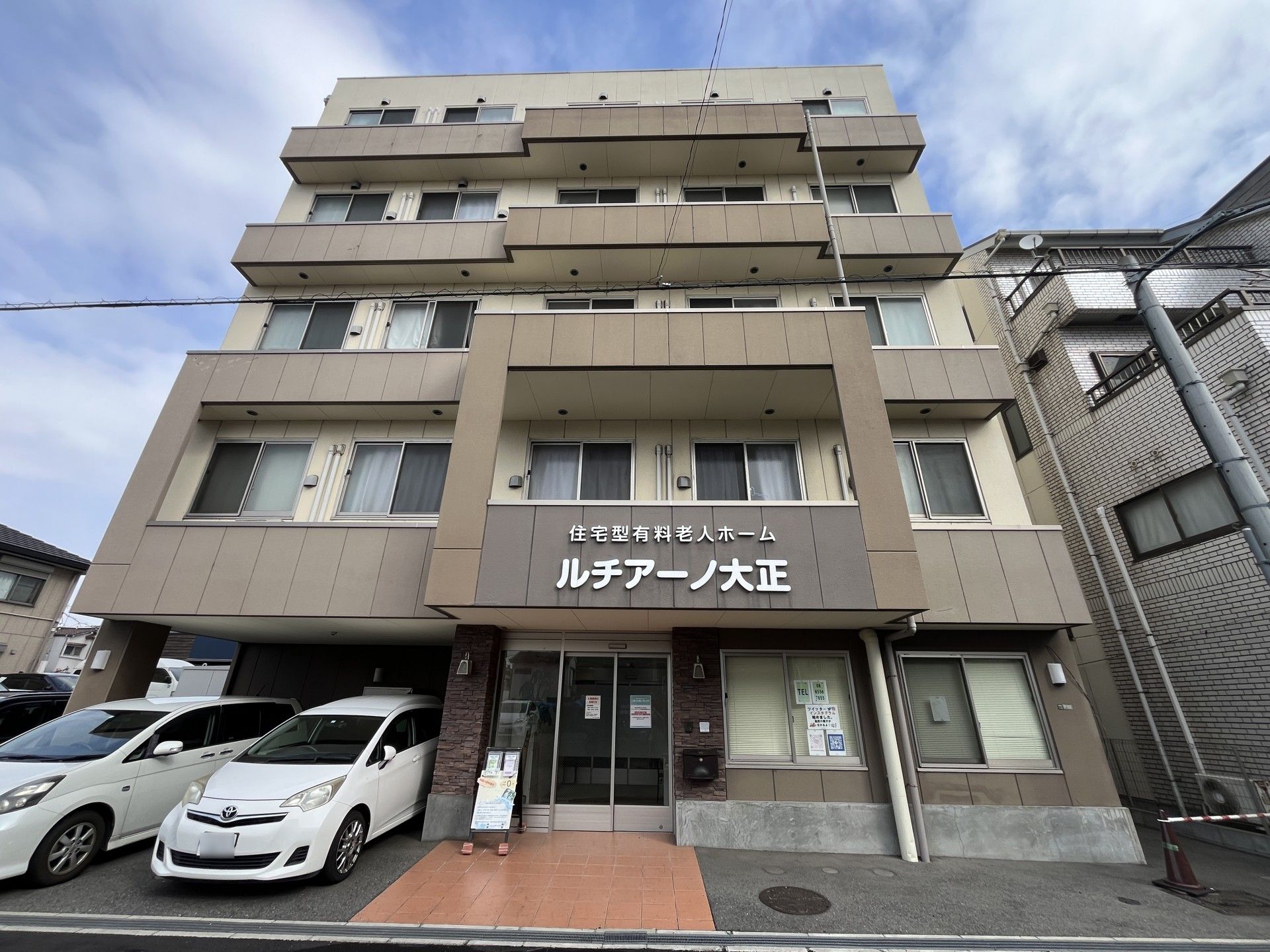 玄関前にはウェルカムボードが置かれ、施設のスタッフ募集の旨と連絡先の添えられた張り紙が貼られている。