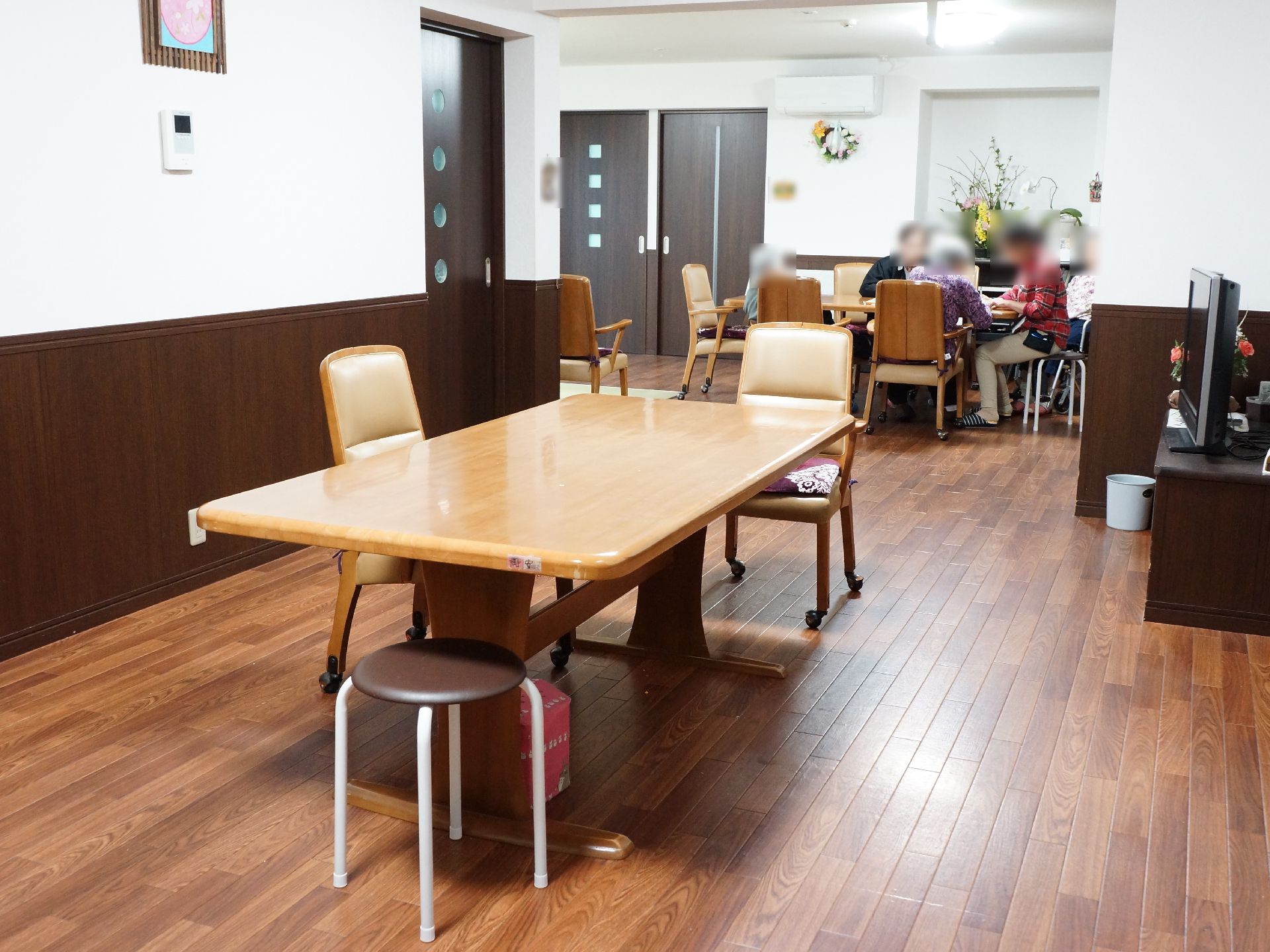奥のテーブルでは、入所者が数名談話している。その近くには、花が活けられていて雰囲気がよい。机には人数に合わせて椅子を置くことができる。