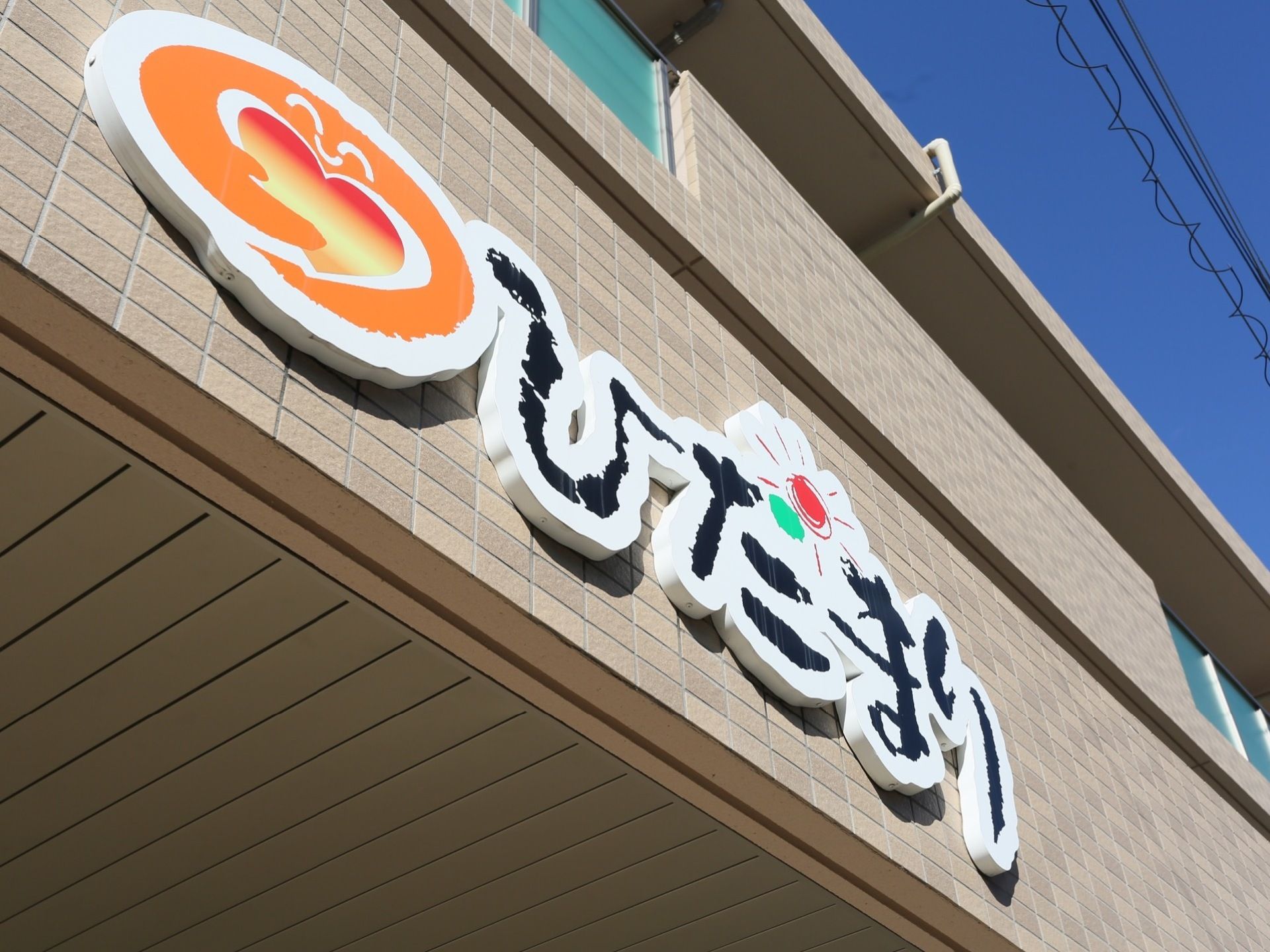 施設の外壁に大きく施設名の看板が設置されている。あたたかみのあるオレンジ色のハートや赤色の太陽のマークも描かれている。