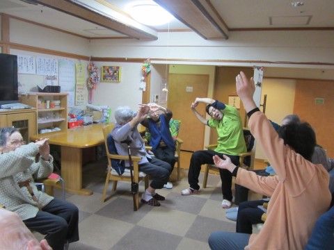 椅子に座り、職員の真似をして腕を頭の上で組んで体操をする入居者たち