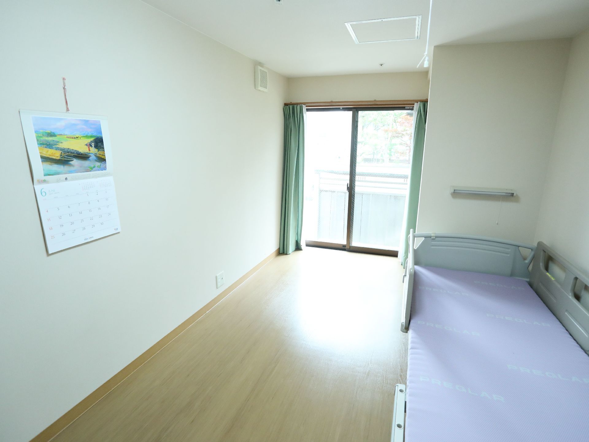 居室にはベッドが置かれている。窓にかかったカーテンは開けられている。壁にはカレンダーがかけられている。