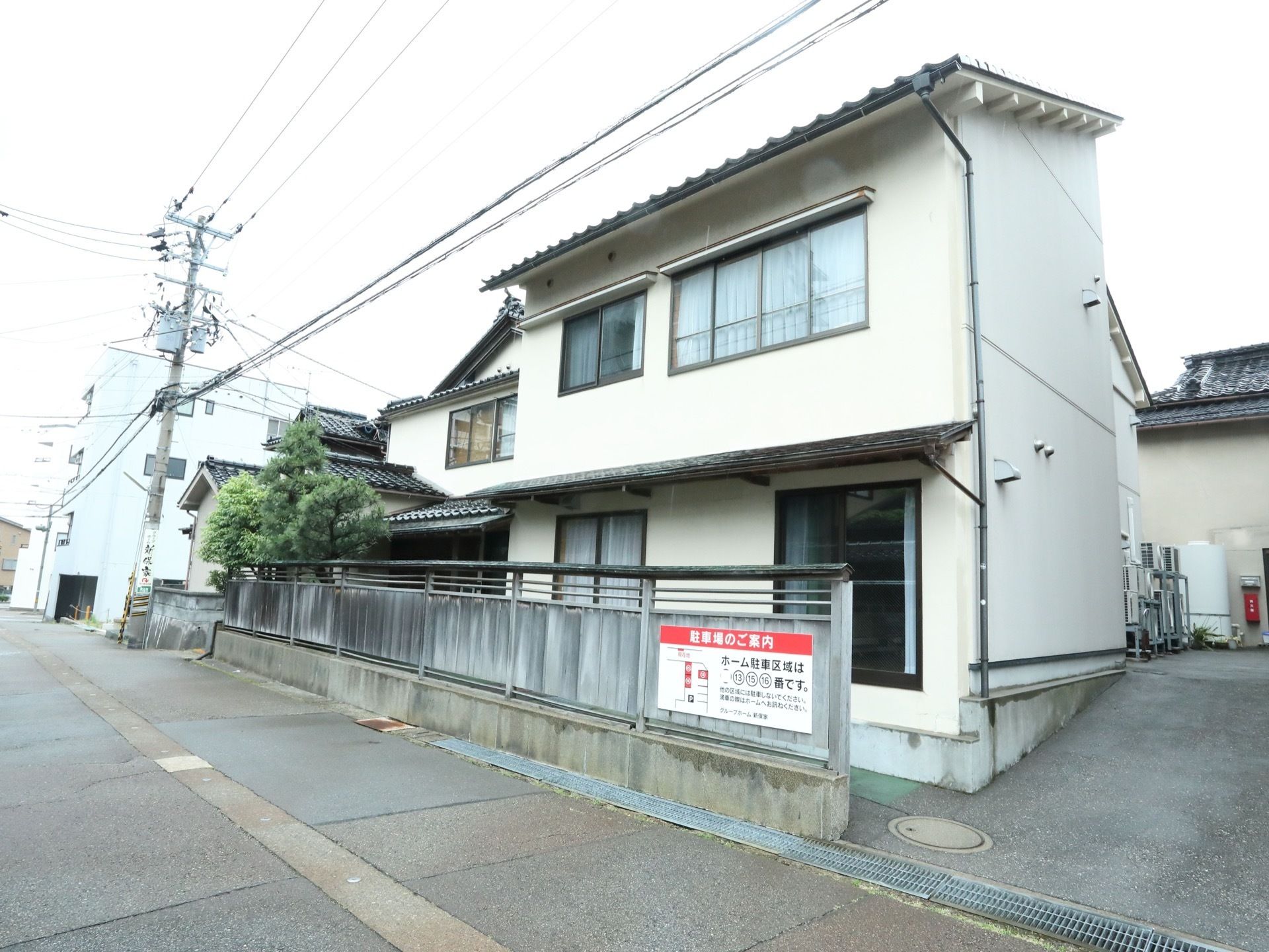 施設は2階建てで白い外観である。施設には塀が設置されている。そこには施設利用者専用の駐車場の案内板が貼られている。
