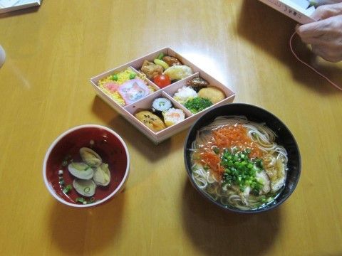 テーブルの上にある、具だくさんの蕎麦と菱形の弁当とアサリのお吸い物