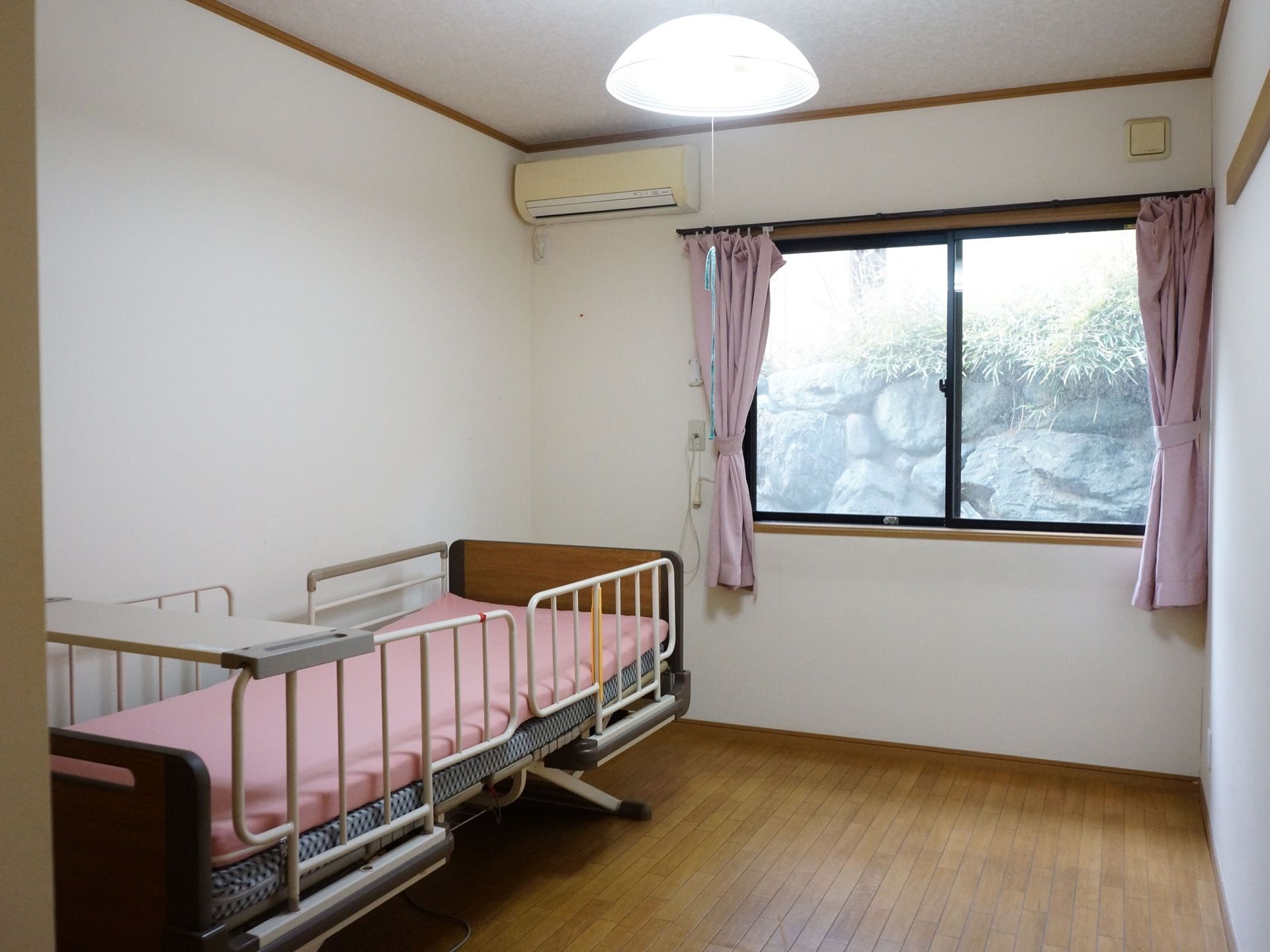 窓から日差しが差し込む、フローリング張りの洋室。机付きの可動式のベッドが置かれている。また、エアコンが完備されている。