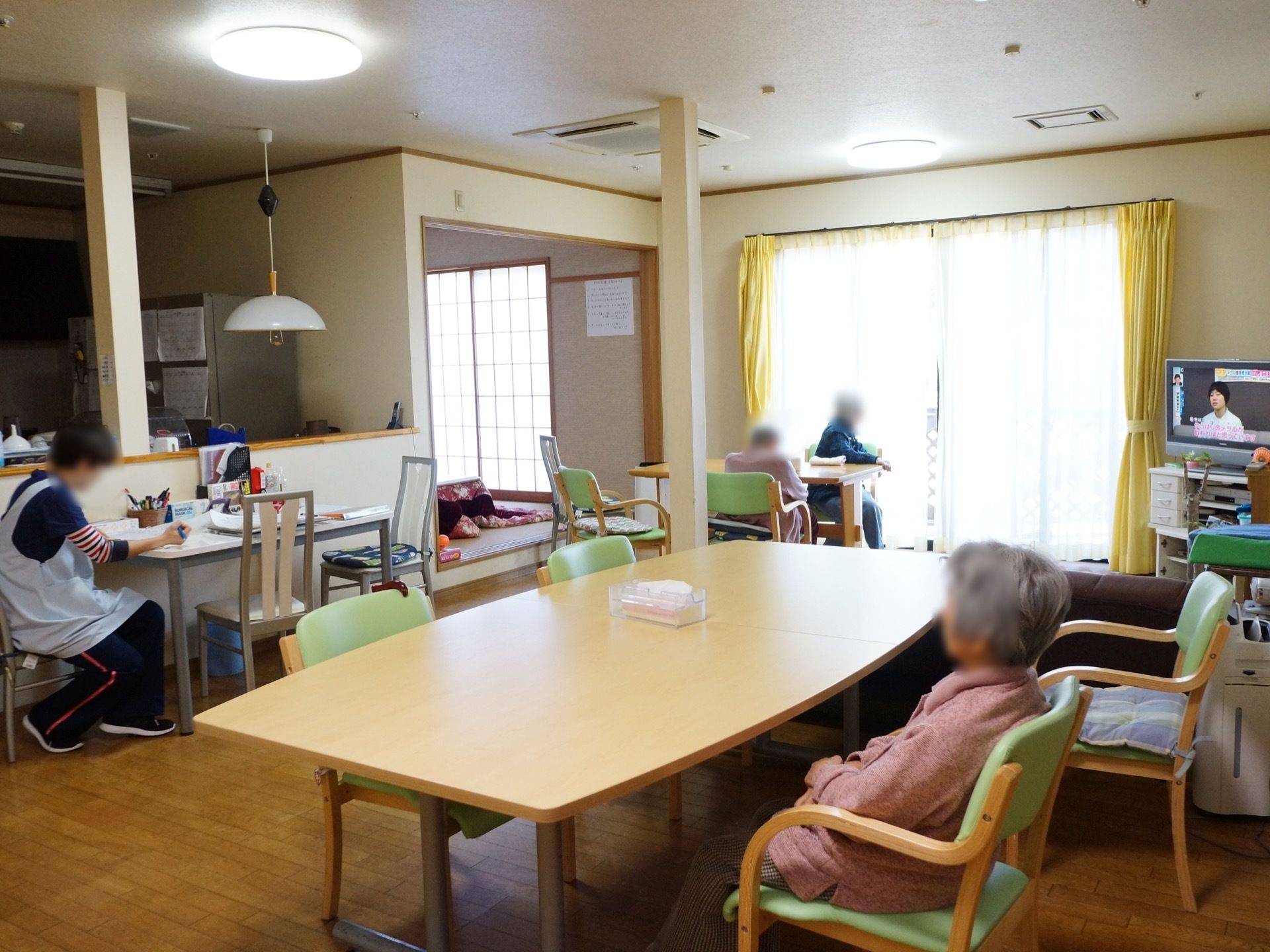 窓から日射しが入り、明るい雰囲気である。テーブルと椅子、テレビが設置されている。部屋の中では入居者様がくつろぎ、スタッフが仕事をしている。