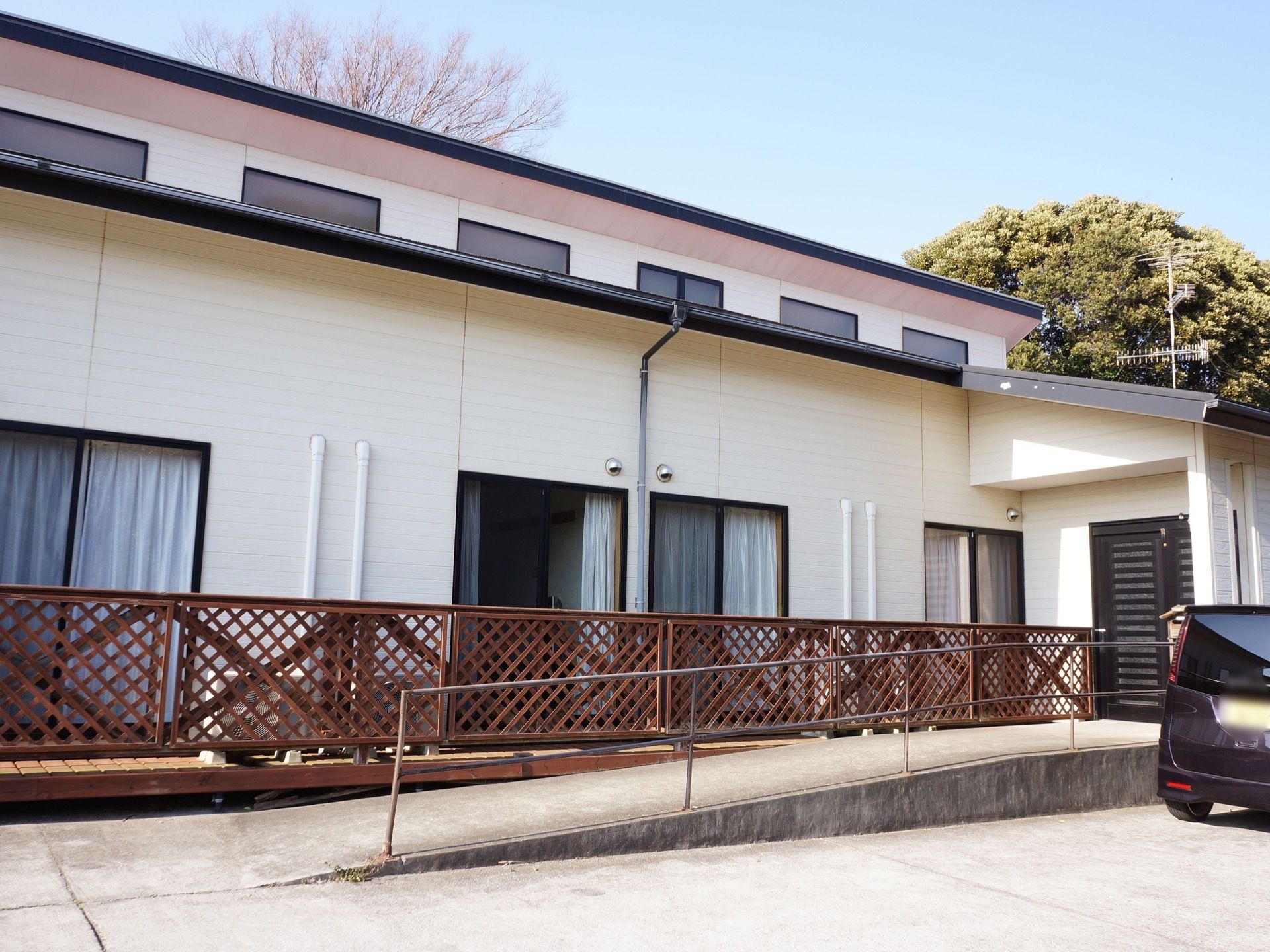 施設は２階建てである。駐車場に面した方にたくさんの窓が設置されている。施設の向こう側に大きな木々が見える。
