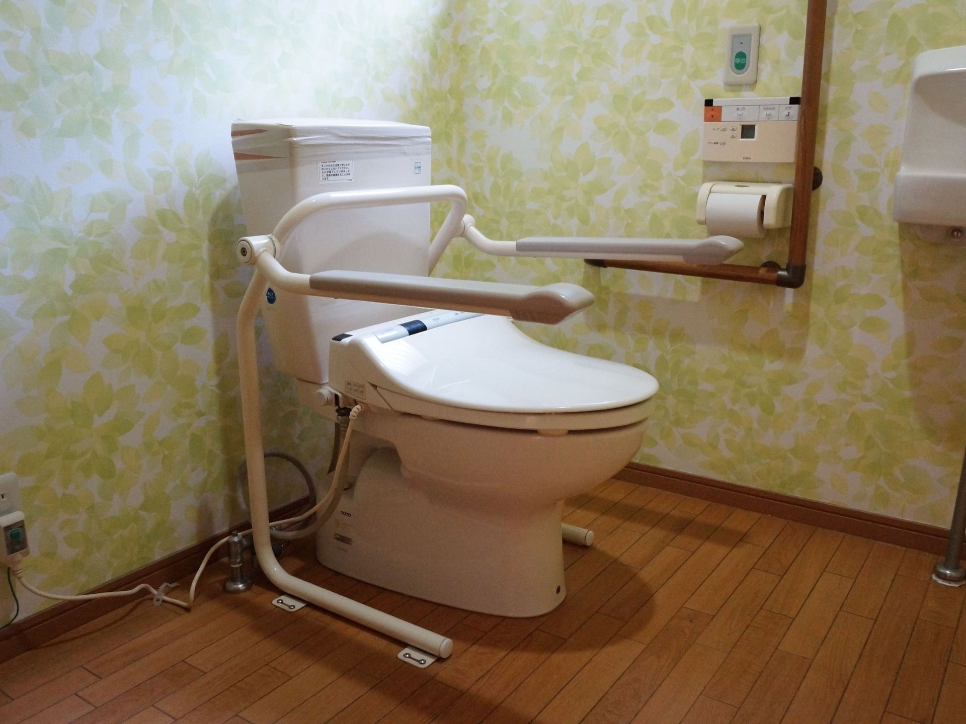 温水洗浄便座のトイレ。壁にはL字型の手すりが付いていて、便器には可動式の手すりが両側に付いている。また、緊急通報装置が設置されている。