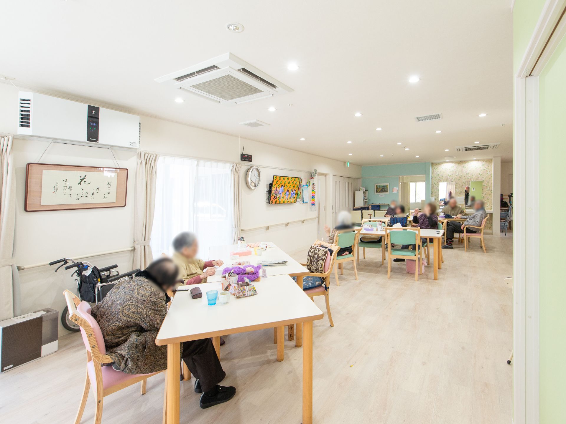 広い部屋にゆったりとテーブルセットが配置され、天井には空調機とスプリンクラーが設置されている。入居者様達が団らんを楽しんでいる。