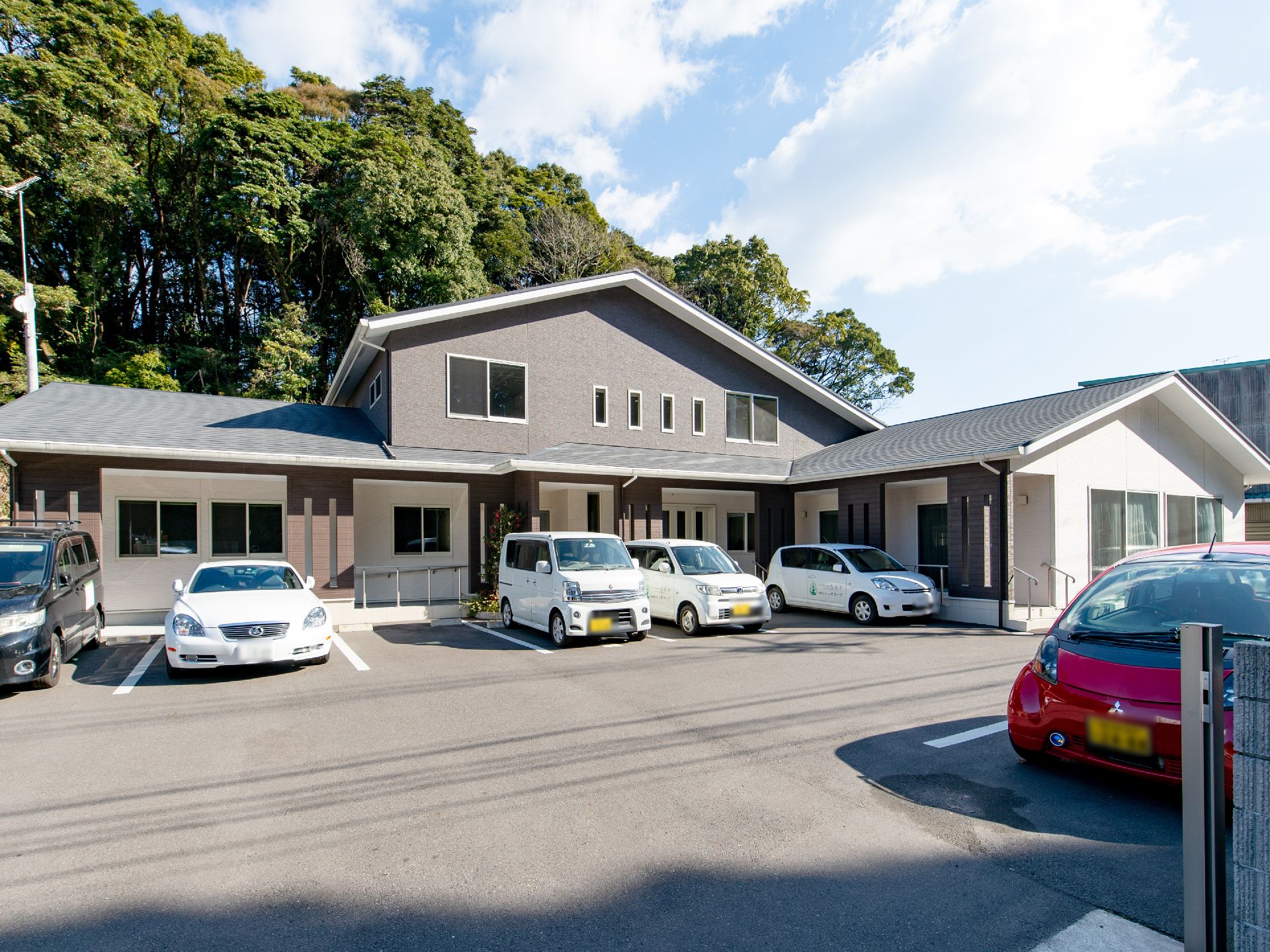 緑の大木をバックに中央が2階建てのグレーのおしゃれな建物である。施設前の通路には手すりがつき、駐車場に車が数台停まっている。