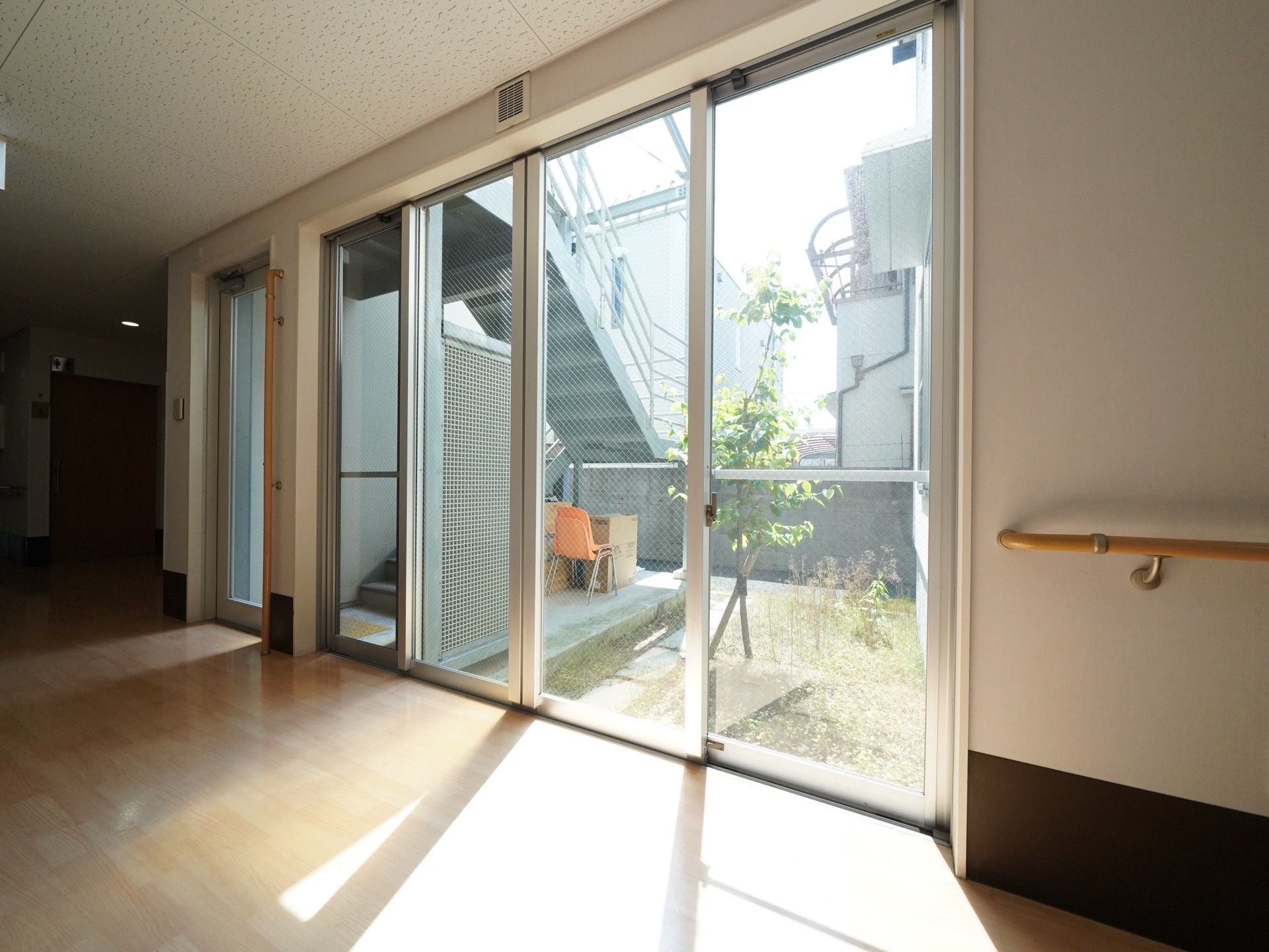 施設内の廊下に設置されたガラスサッシ扉からは、日差しがたっぷり入り込み、外の中庭の木々が見えている。
