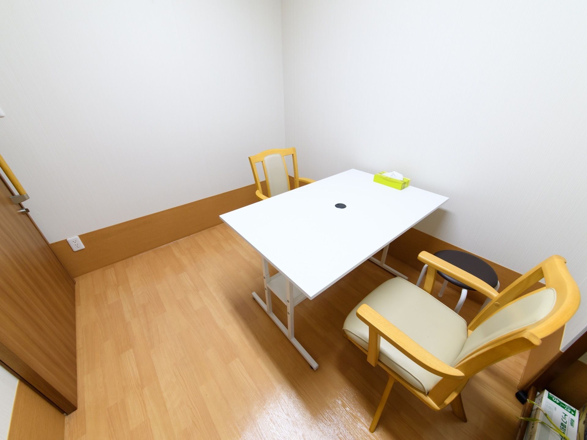 白いテーブルと、椅子が２脚向かい合うような形で設置されている。奥に背もたれのない椅子も置いてある。テーブルの上にはティッシュが置いてある。