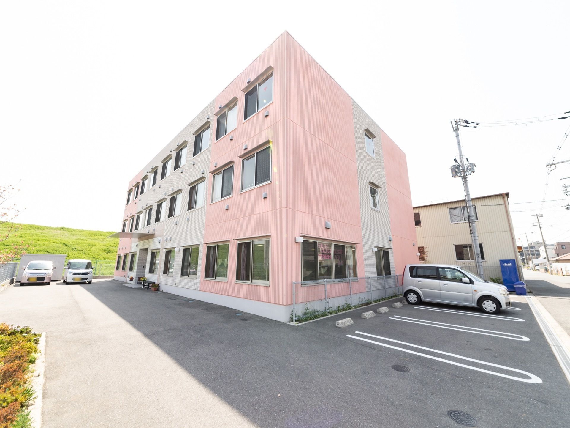 ３階建ての建物は、ピンクと薄いグレーに塗り分けられている。建物の横は大和川の土手になっている。駐車スペースが用意されている。