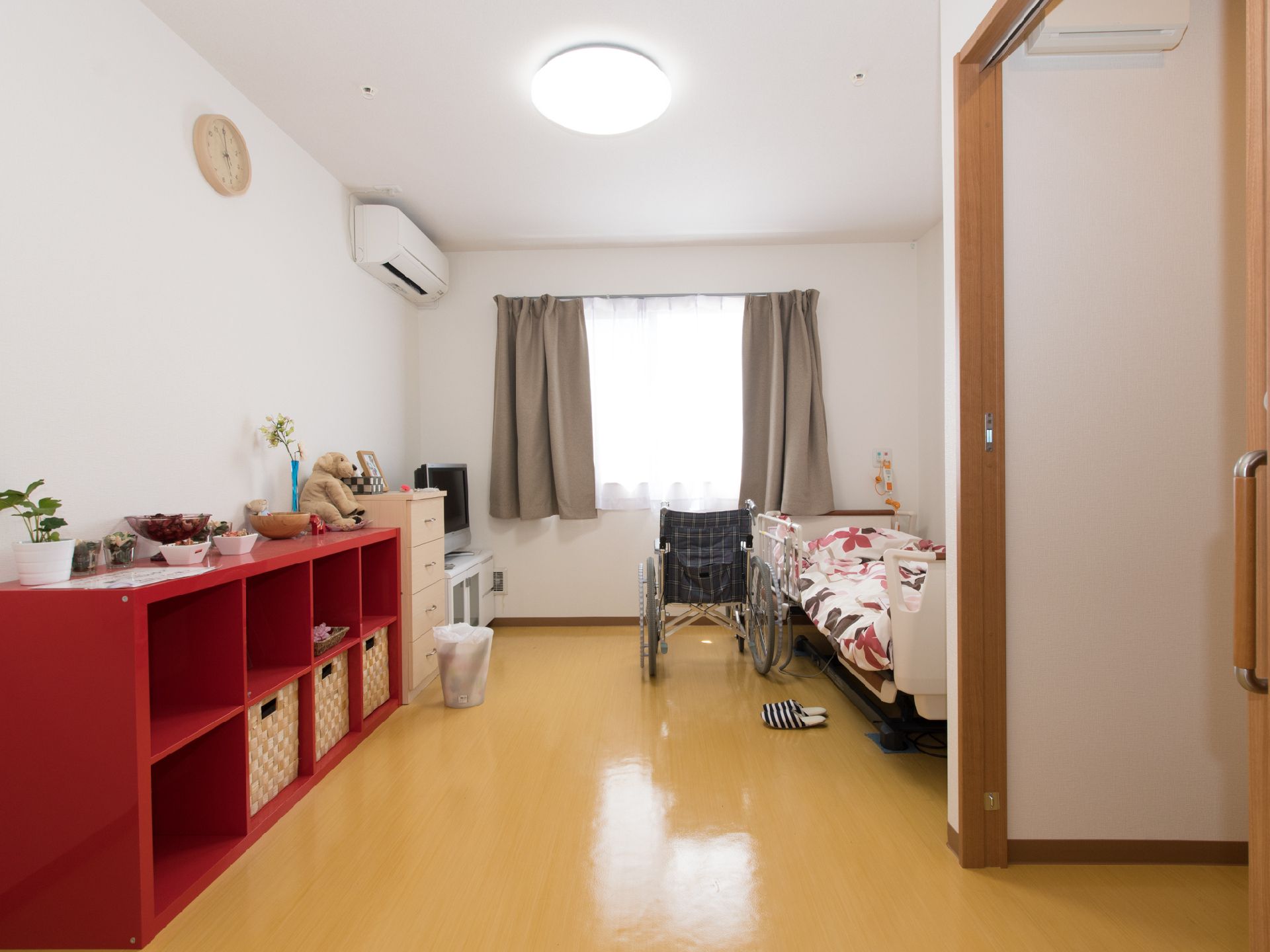 赤いチェストが壁沿いに設置され、車椅子がベッドの脇に置かれた明るい部屋