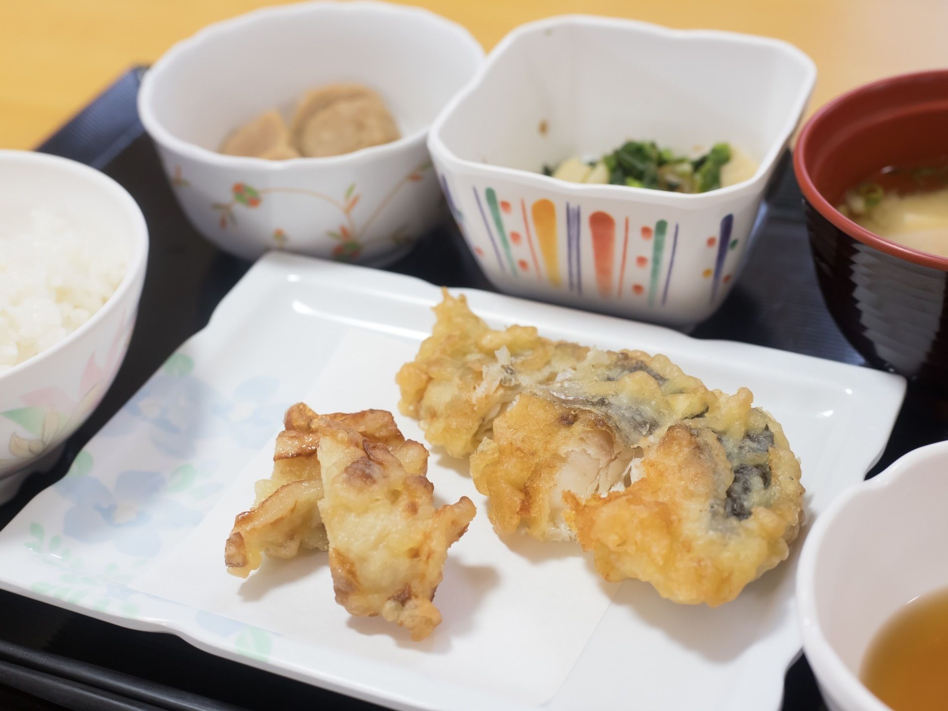 プラスチックの食器が使われており、主菜のほかに副菜が２つ、味噌汁が用意されている。この日は魚の天ぷらで、天つゆもお盆に乗っている。