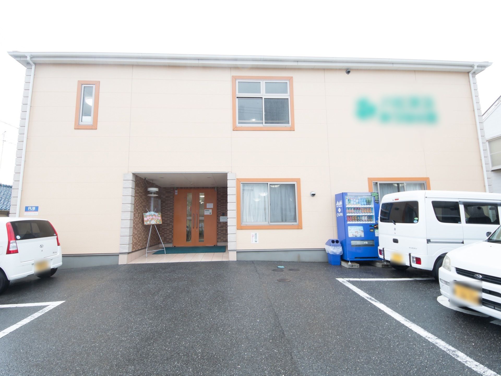 オフホワイトの外壁にブラウンの枠の窓が並んだ２階建ての外観である。建物傍に自動販売機を備え、前方に駐車スペースを設けている。