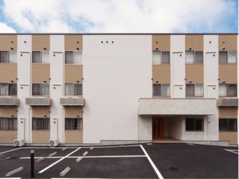 白とベージュ２色が使われた外壁のモダンな雰囲気の３階建て建物である。施設の前には広い駐車場を設置している。