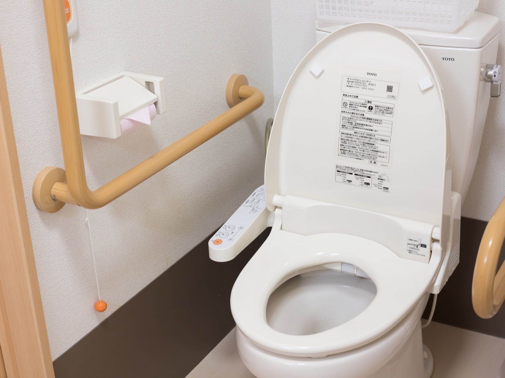 白くきれいに磨かれた、便座カバー付きのトイレがある。側面の壁には手すりやつかまり用の棒が備え付けられている。