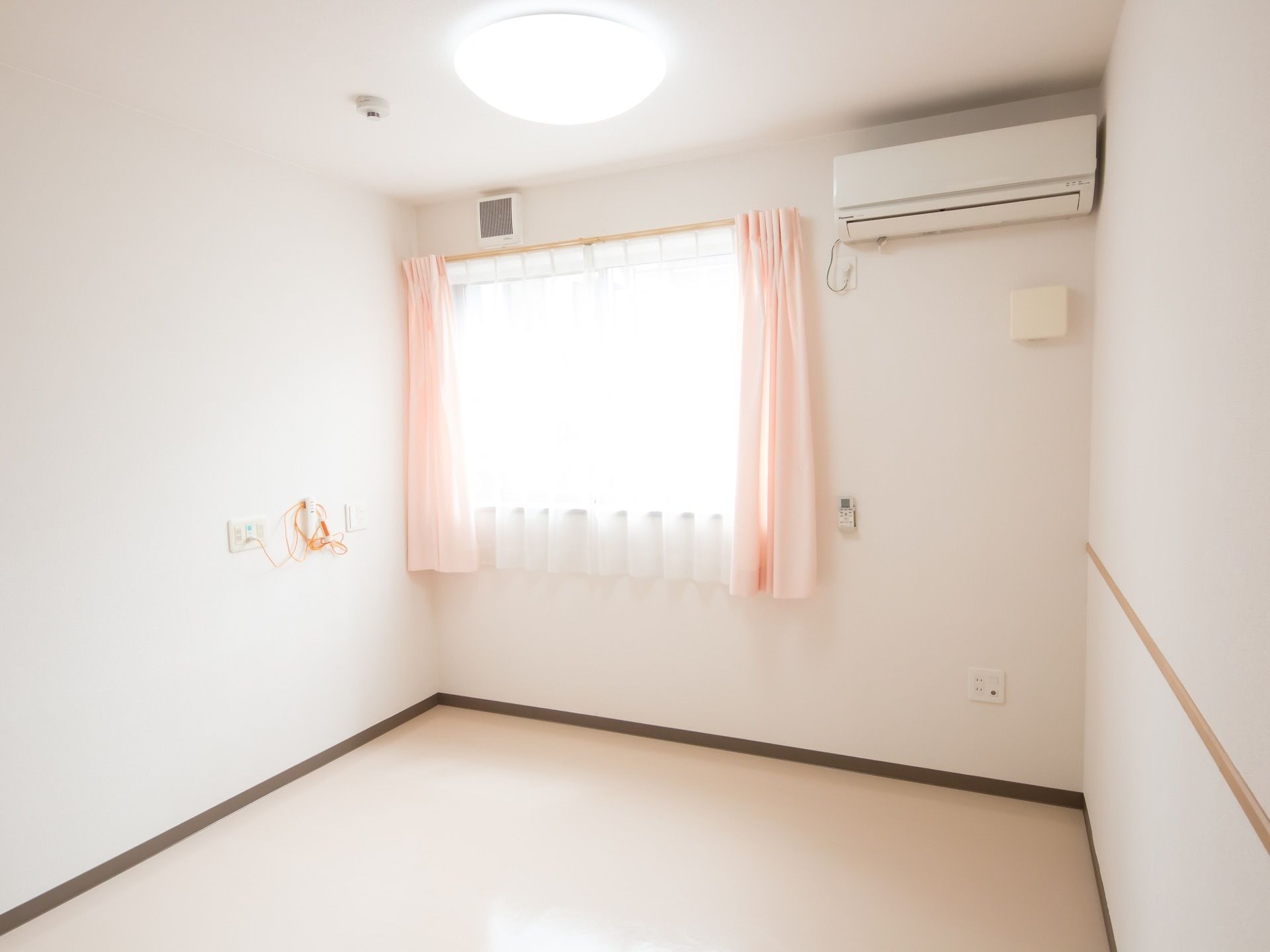 居室の天井近くの壁にはエアコンが取り付けられている。また、奥の壁には窓が設置されており、桃色の明るいカーテンがかけられている。
