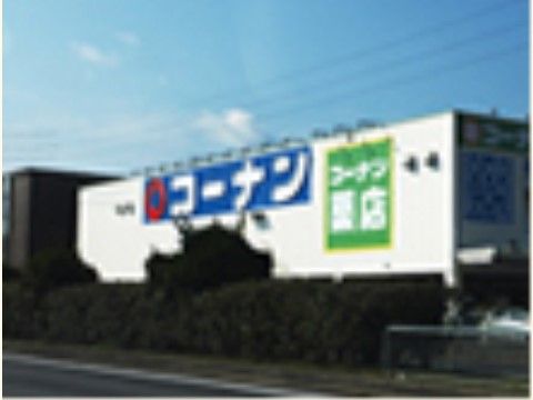 道路沿いにある、緑の木々で囲われた白い横長の建物。壁面上部には青いベースに白文字で、コーナンと書かれた大きな看板が掲げられている。