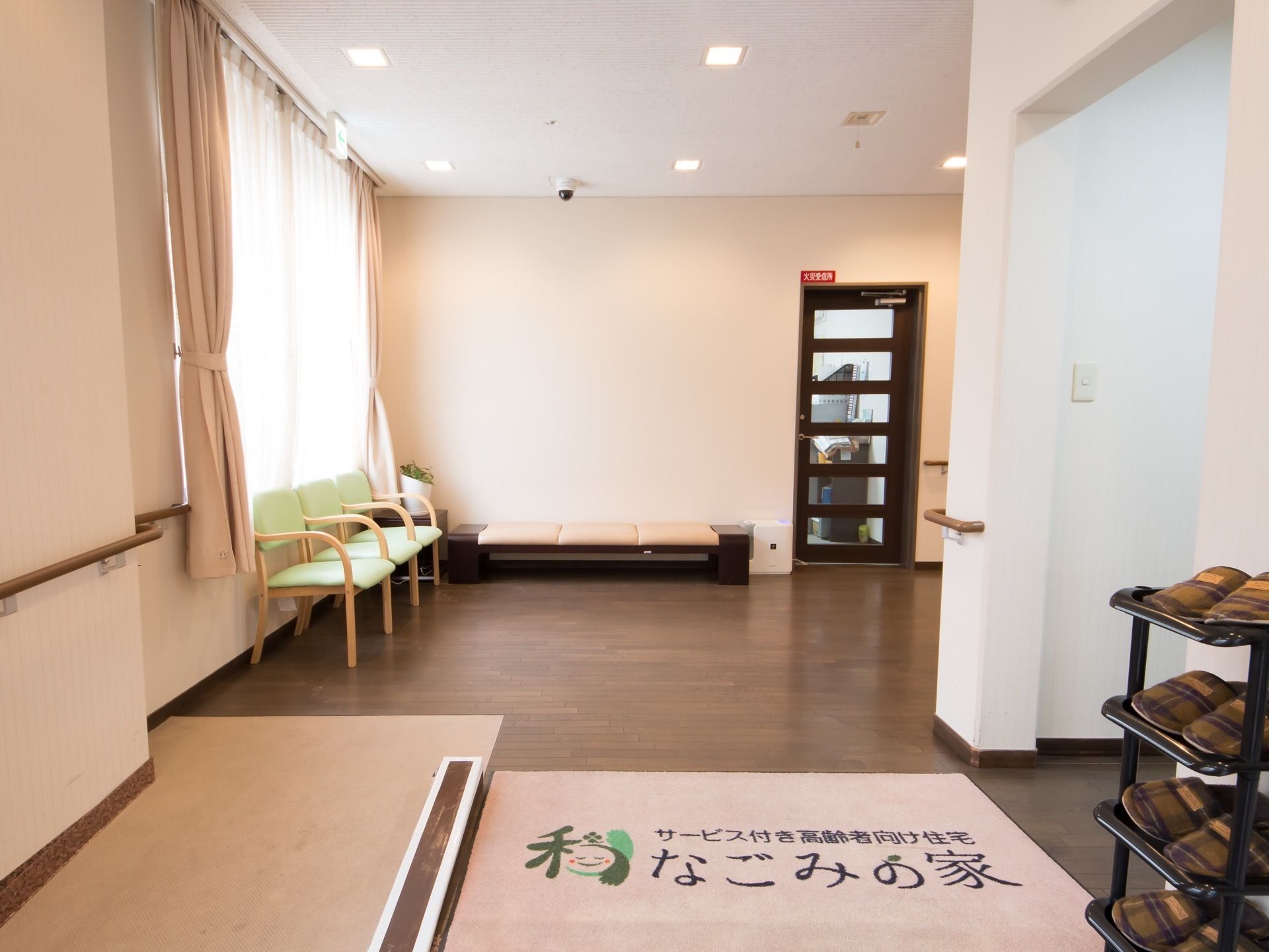 玄関には右にスリッパの並んだ棚、奥には椅子とベンチが配置されている。ガラス張りのドアも見える。天井には防犯カメラが設置されている。