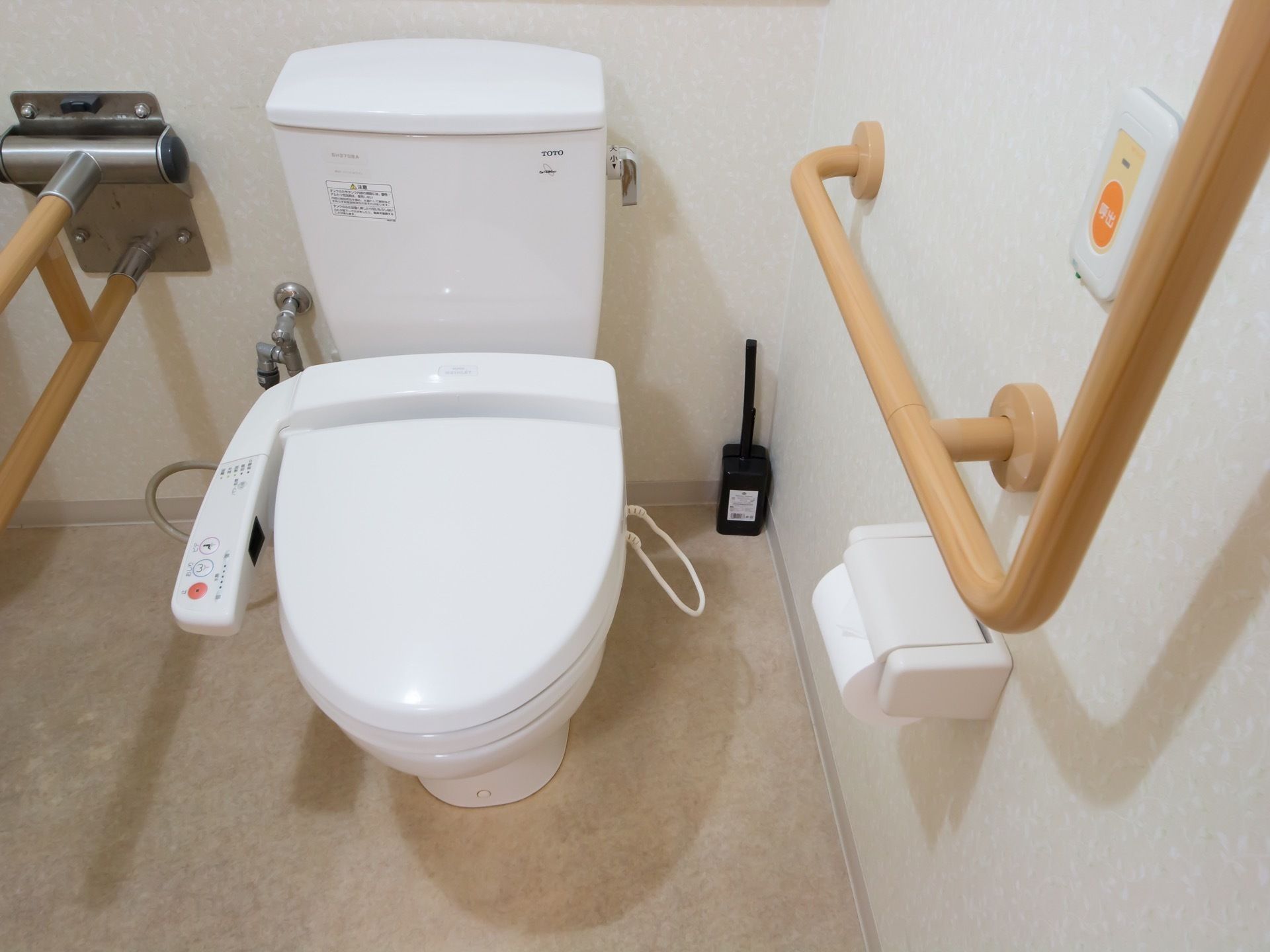 洋式便器の左右には、ベージュの手すりが設置されている。トイレは温水洗浄便座である。壁には緊急呼出しボタンがある。