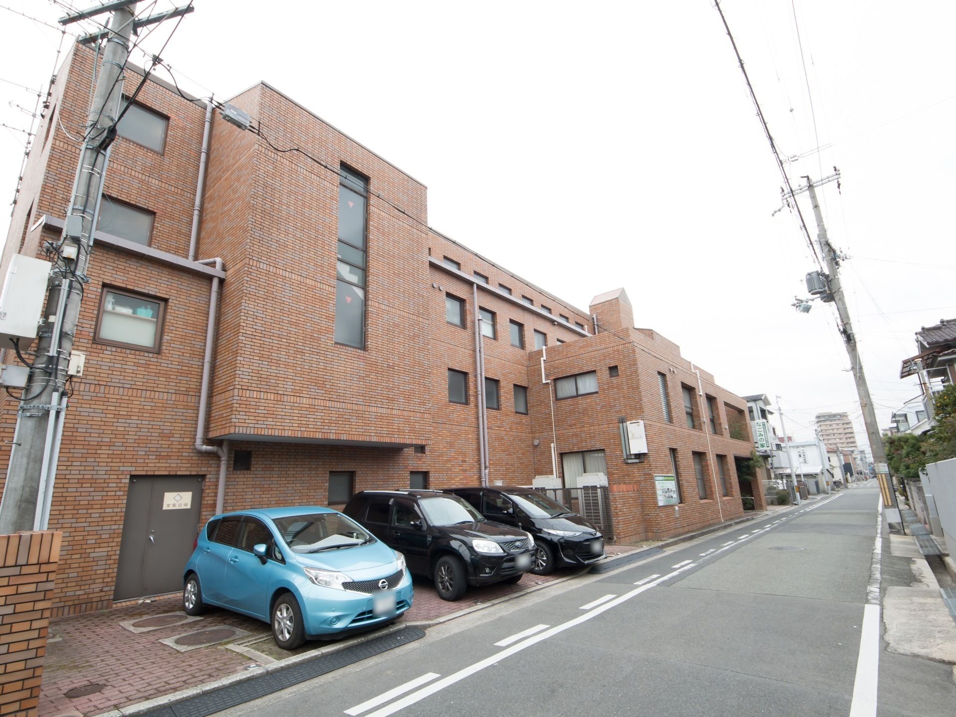 茶色の４階建ての建物。手前部分は２階だが、奥に向けてフロアが増えて行く構造になっている。手前には４台ほどの駐車スペースが確保されている。
