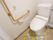 温水洗浄便座の洋式トイレが設置されている。左の壁にはＬ字型の手すりがあり、緊急コールも備えつけてある。