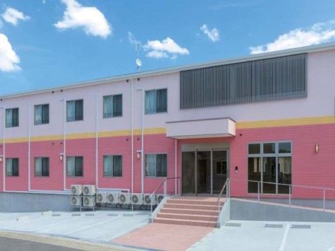中央に黄色のライン、上は薄いピンク、下は濃いピンクの外装の２階建ての建物。入り口の正面には階段があり、建物に沿ってスロープが設置されている。