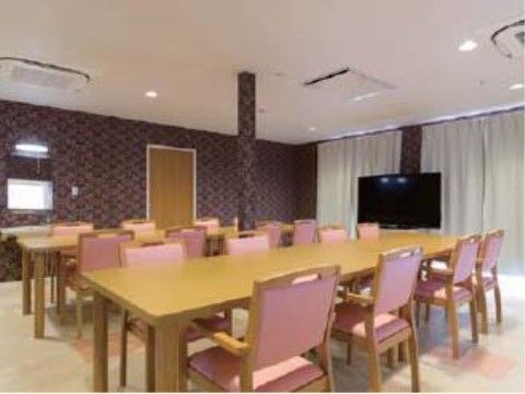 柄模様の濃い色の壁紙に、長テーブルが２卓、ピンク色の椅子が並んだ室内。窓際には大画面テレビが置かれている。