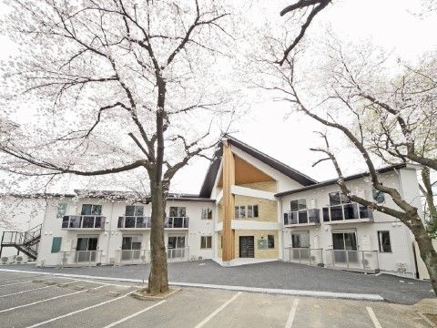 施設の目の前に駐車スペースがあり、一部に桜の木が植えられている。白を基調としたおしゃれな外観の建物。