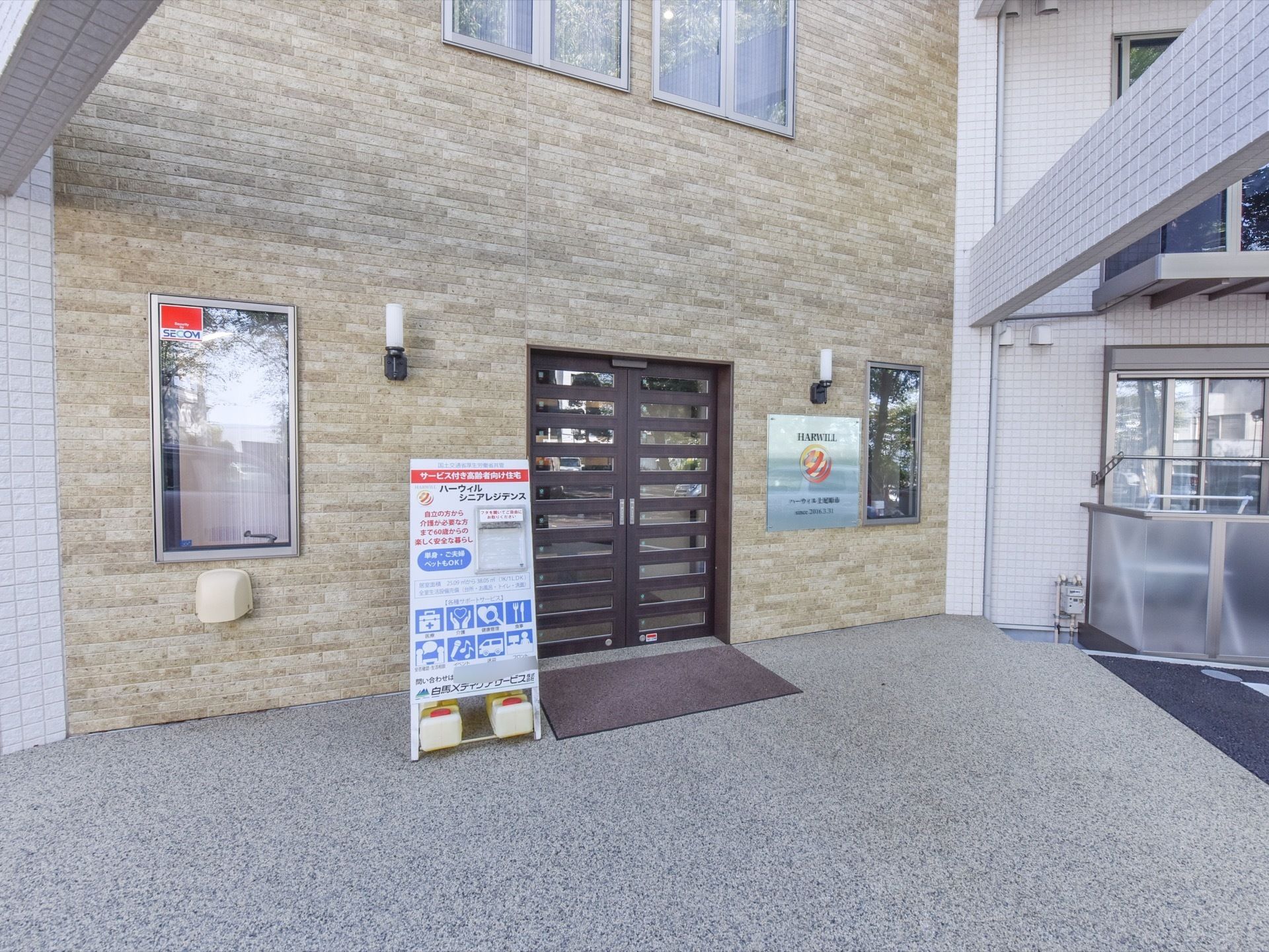 玄関ドアがある部分はベージュ色、その横につながる部分は白色の壁になった建物である。ドアの右には施設名が貼られ、左には看板が立てられている。
