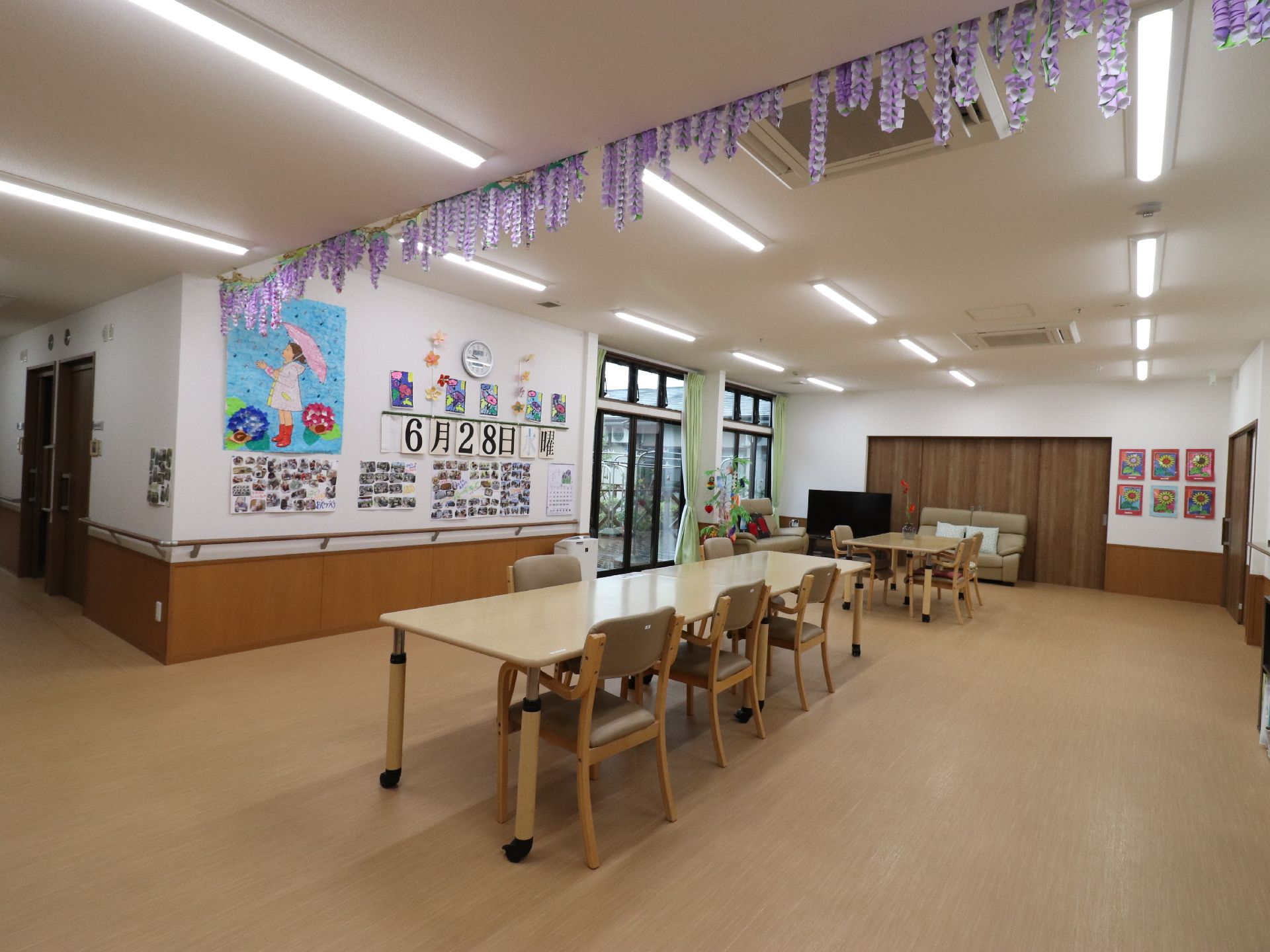 左側には今日の日付と折り紙の貼り絵が飾られている。窓も大きく取られているので明るく感じる。奥にはテーブルとテレビがある。