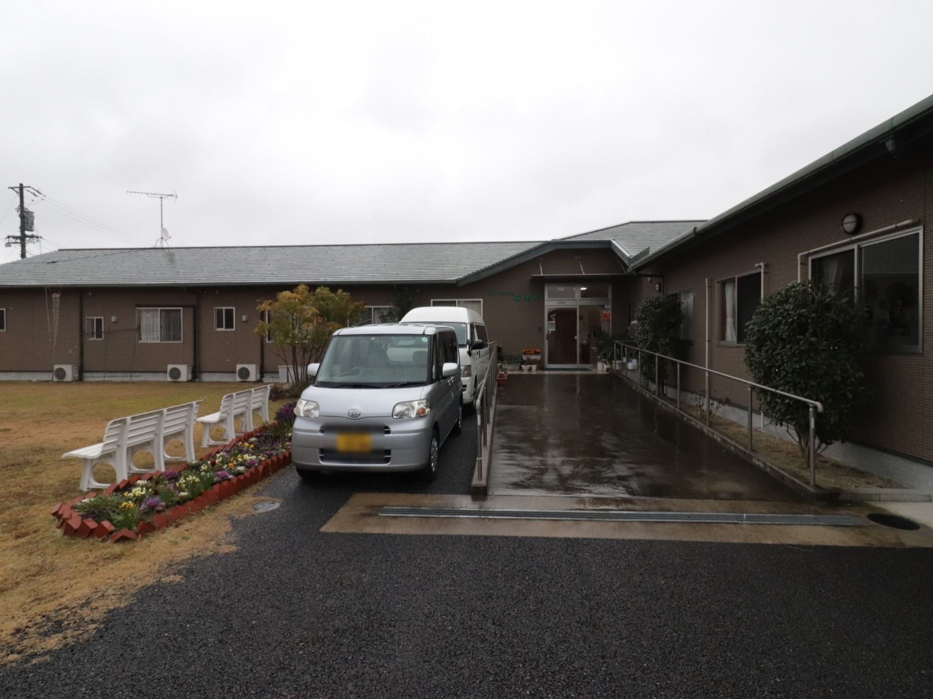 施設玄関前には手すりが付いたスロープがある。スロープの横には車を止められるようになっている。車の近くには花壇とベンチがある。